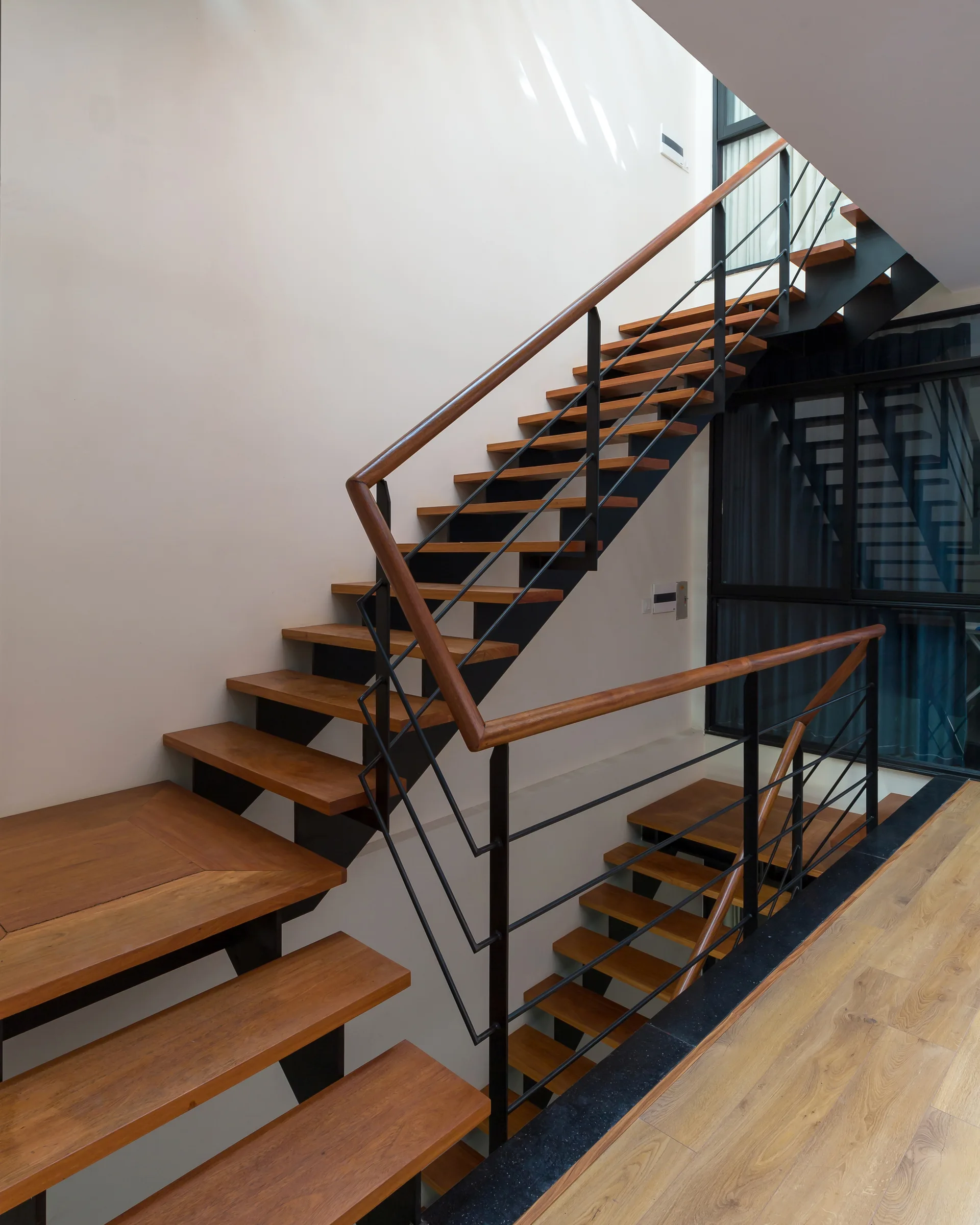 An open-riser wooden staircase features warm-toned treads and a black metal handrail against a plain off-white wall.