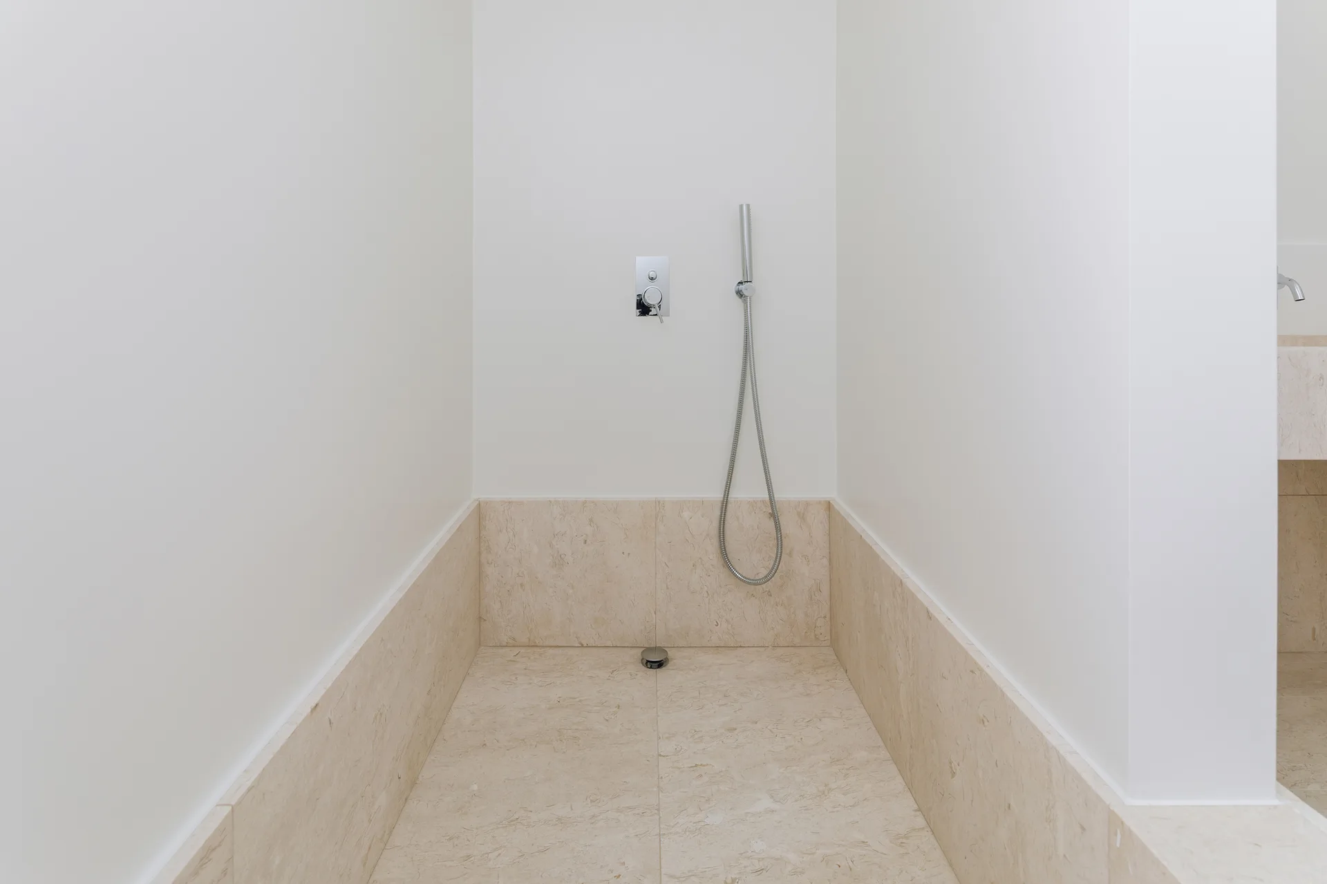 A minimalist shower alcove features beige natural stone floor tiles and half-height wall cladding paired with a chrome handheld sprayer.