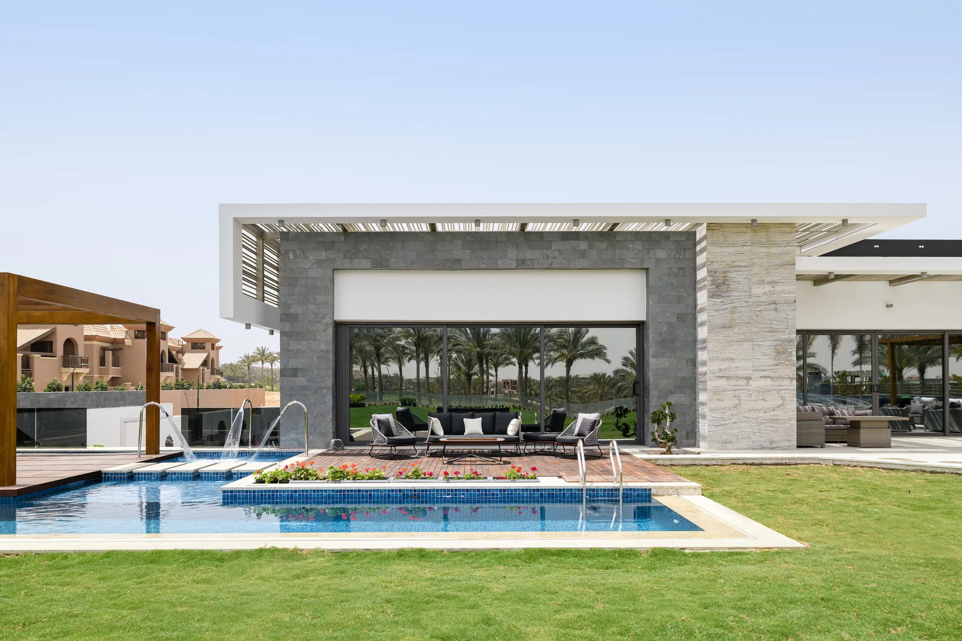 A backyard swimming pool with fountains sits next to a wooden deck featuring dark patio furniture against a grey stone-clad modern home.