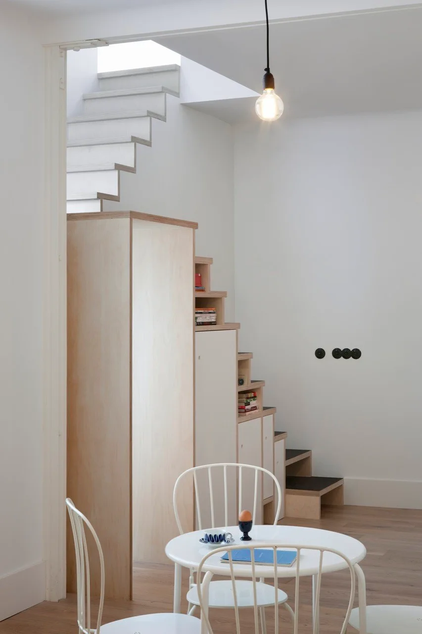 A minimalist white dining area features a round table and chairs next to a light wood plywood staircase with built-in storage cabinets.