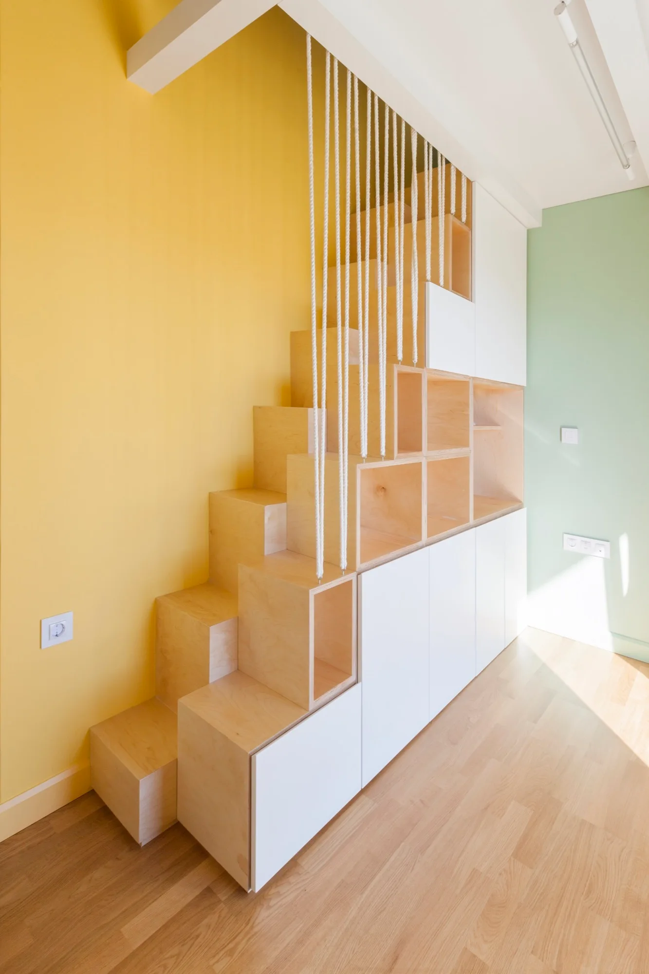 A custom plywood staircase with integrated storage cabinets and open shelving features a vertical white rope railing against a vibrant yellow wall.