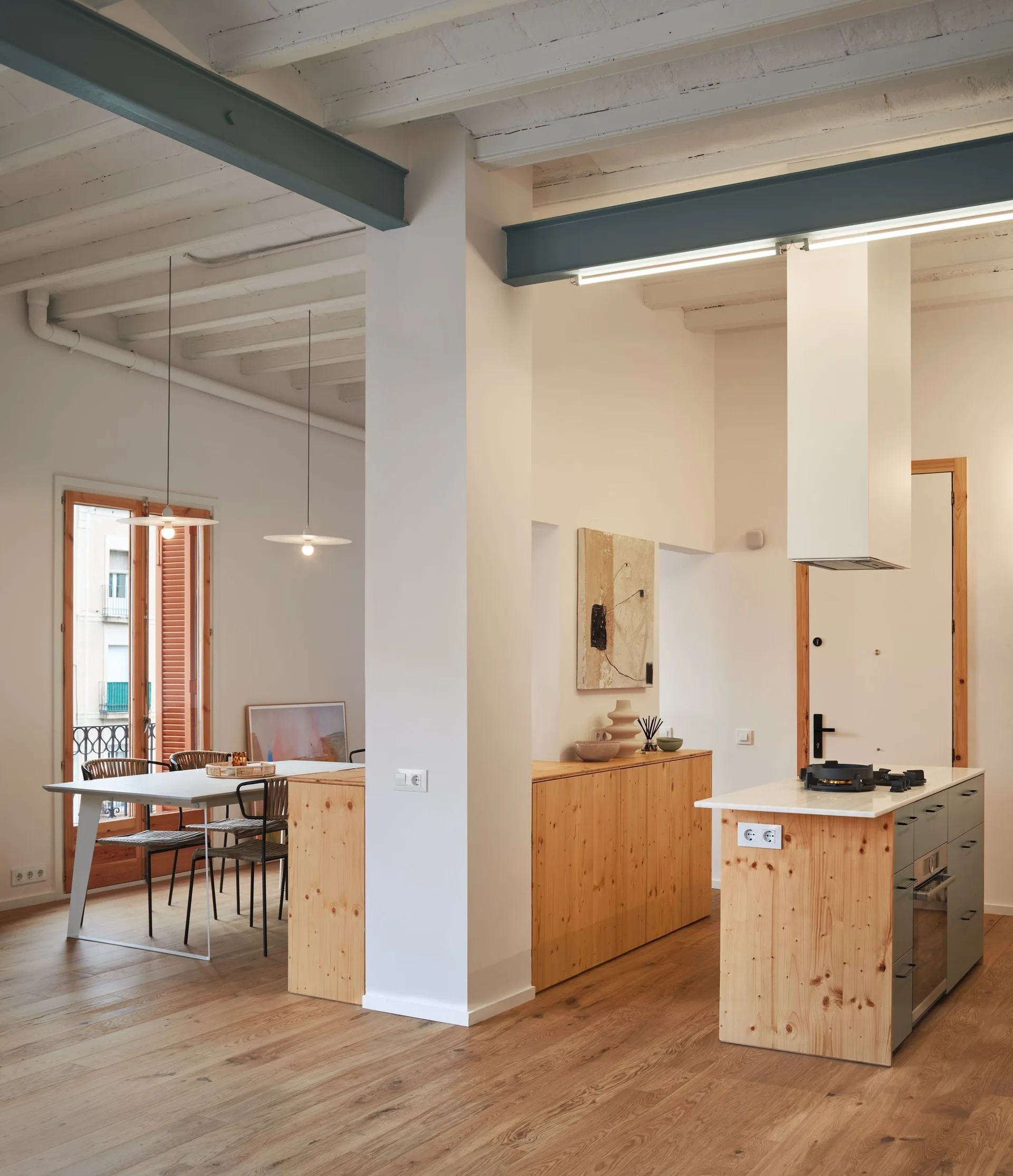 A modern open kitchen and dining area with knotty pine cabinetry, a central island, painted wooden ceiling beams, and a white dining table.