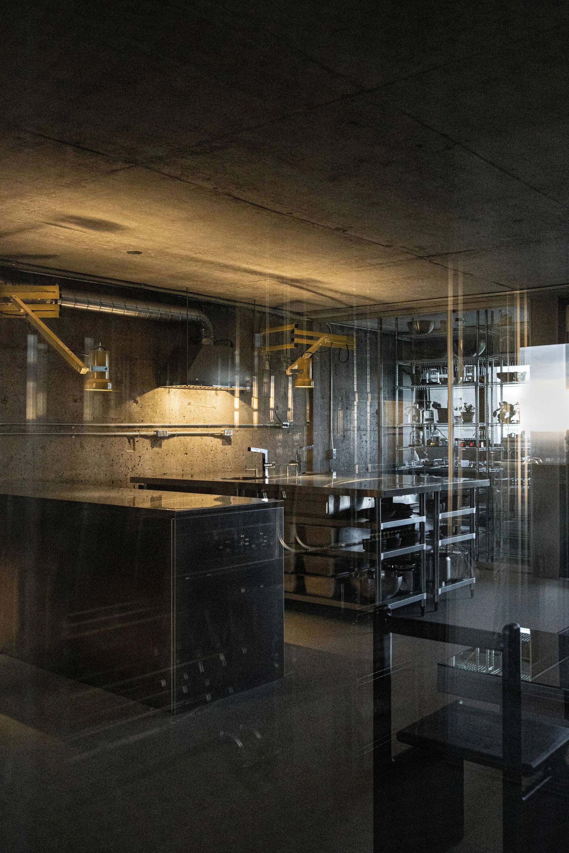 An industrial kitchen features raw concrete walls, a stainless steel island, and yellow scissor-arm wall lamps seen through a glass partition.