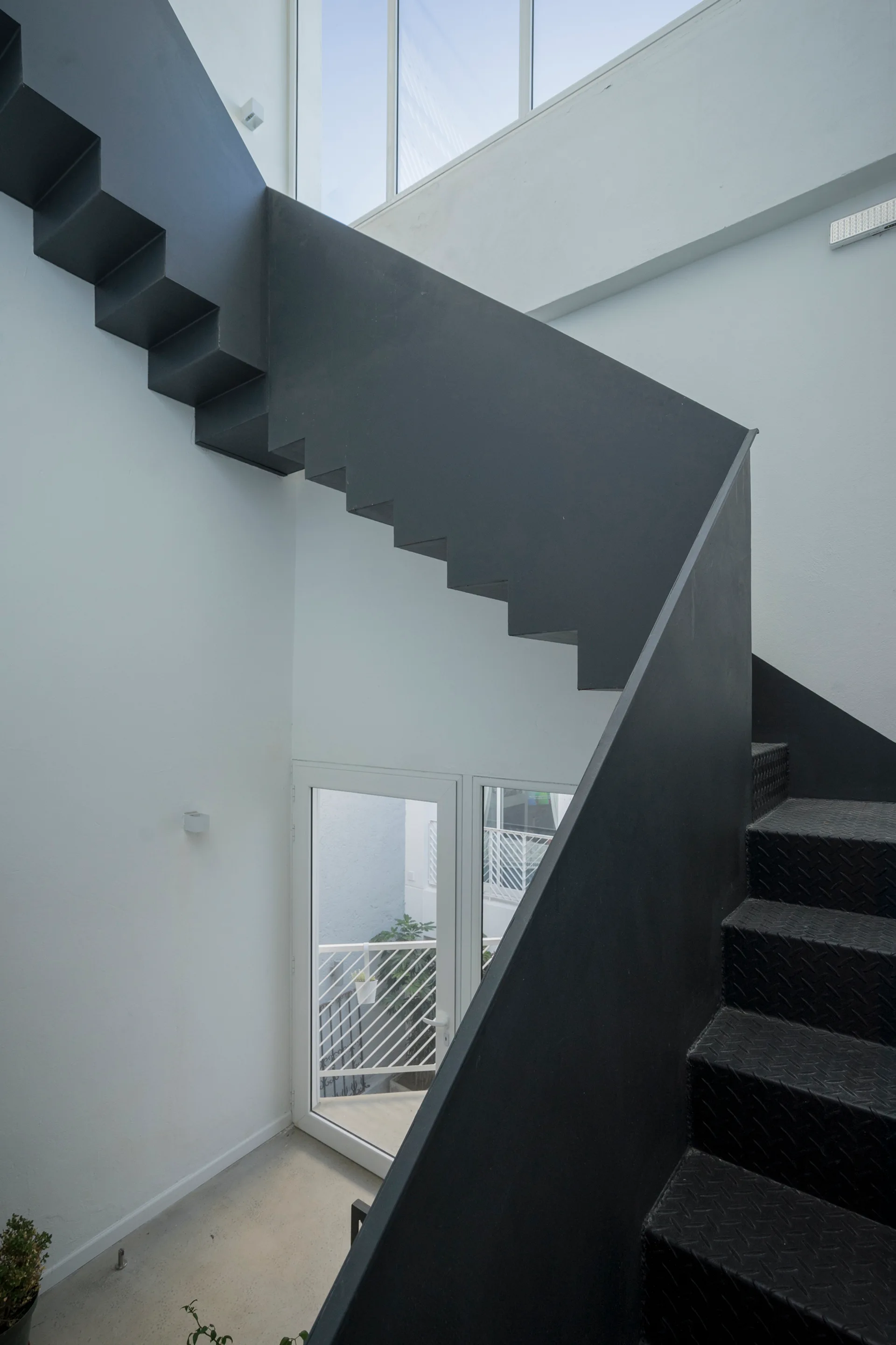 A dark grey metal staircase with a zigzag profile stands against clean white walls in a modern minimalist space with a glass door below.