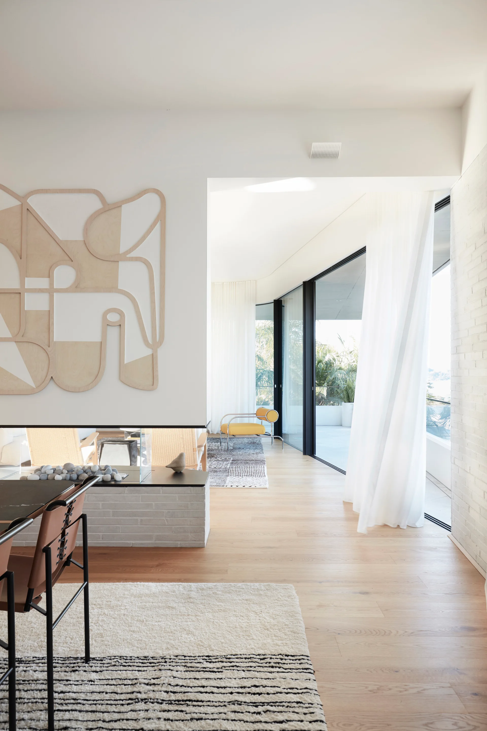 A modern living area features a low-profile white brick fireplace, a large wooden abstract wall sculpture, and sliding glass doors to a terrace.
