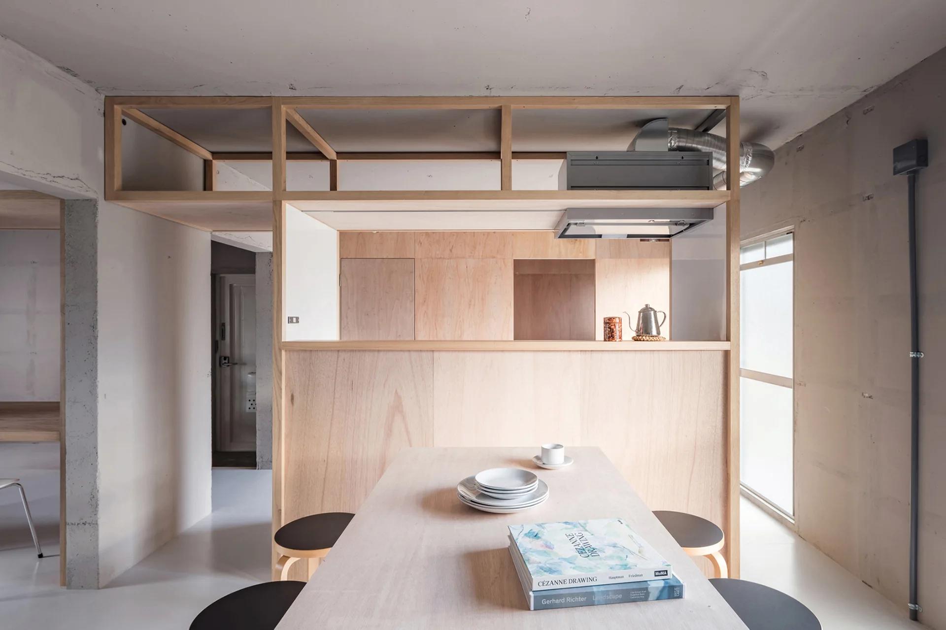 A minimalist dining table and stools sit before a large plywood partition wall under an exposed concrete ceiling.