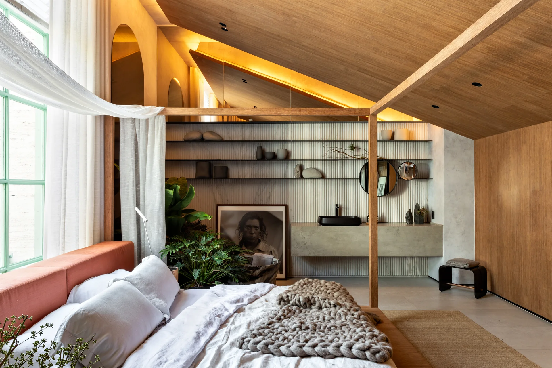 A modern bedroom featuring a wooden canopy bed, chunky knit throw, and a sloped wood ceiling with a vanity area in the background.