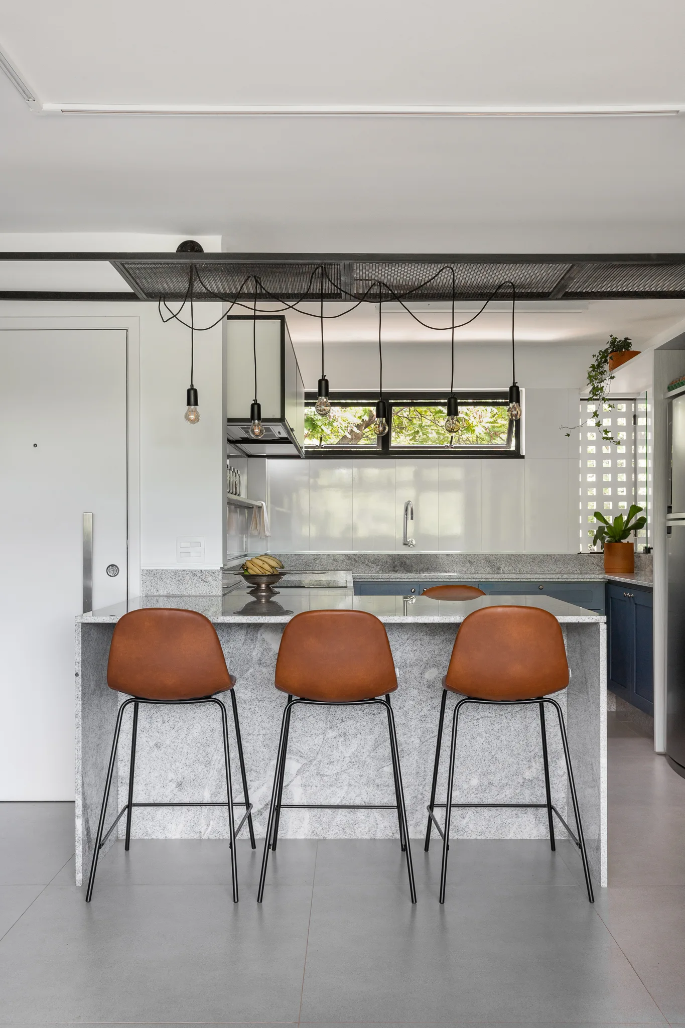 A modern kitchen features a grey stone waterfall island with three tan leather bar stools under industrial exposed bulb pendant lights.