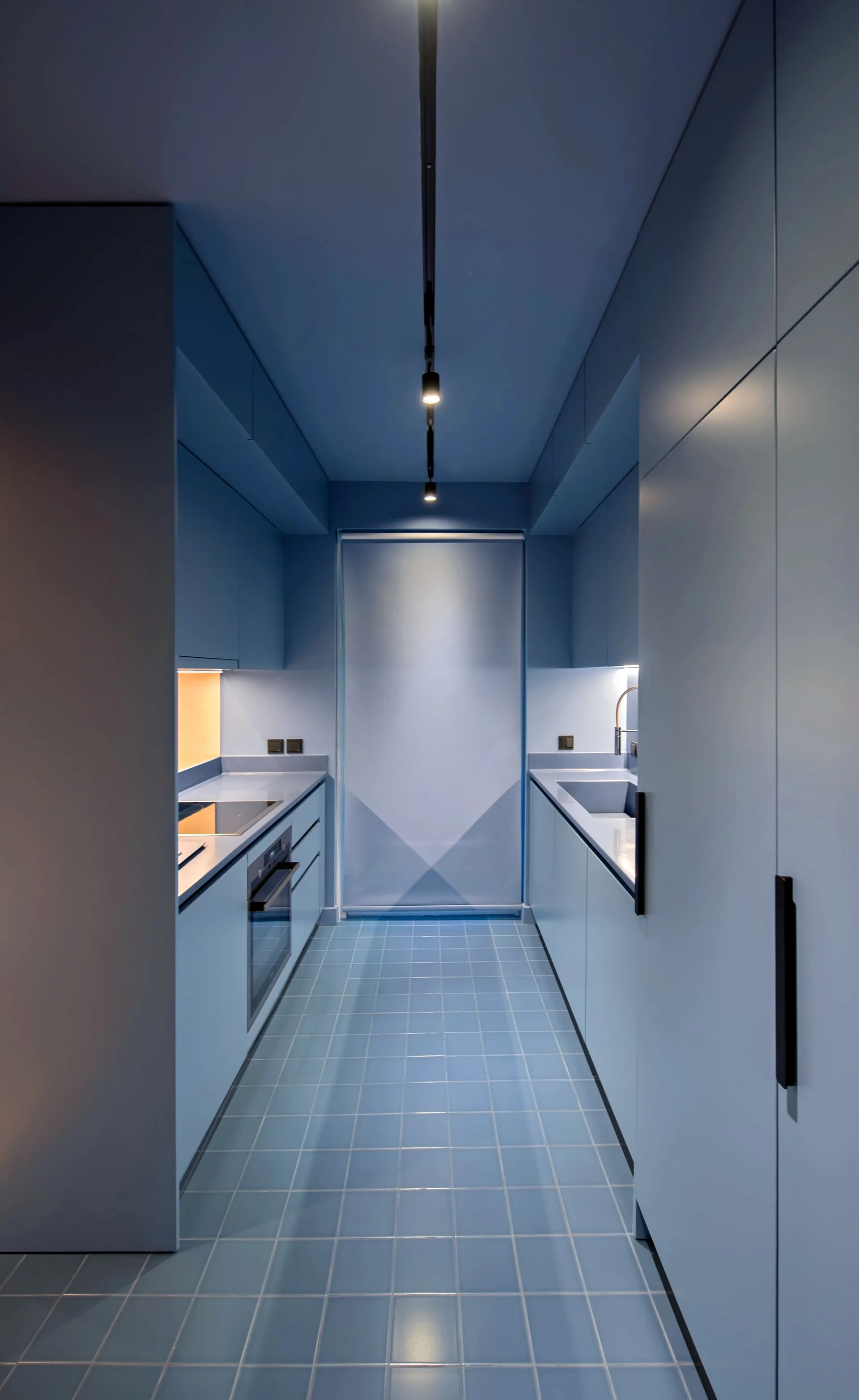 A narrow galley kitchen features monochromatic light blue cabinetry, walls, and matching square floor tiles under a black track light.