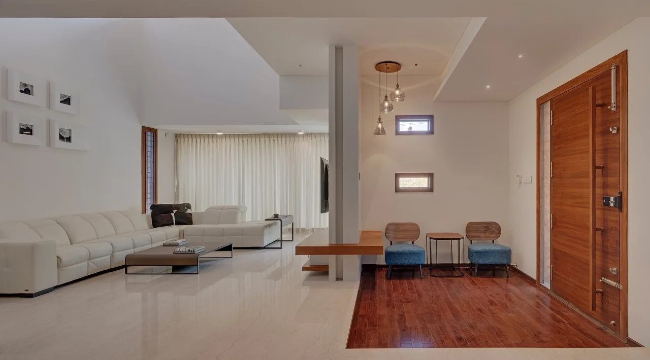 A split-level living room features a white leather sectional sofa on polished stone flooring next to a wood-floored entryway with two blue chairs.