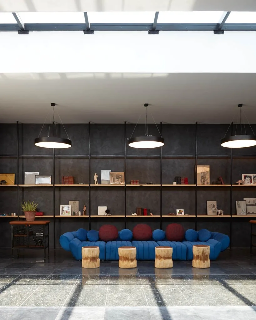 A long blue tufted sofa sits before an expansive black metal shelving unit, complemented by four raw wood log stools under a bright skylight.