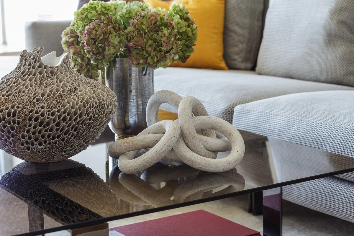 A glass coffee table styled with a textured honeycomb sculpture, stone-like interlocking rings, and a vase of flowers in front of a grey sofa.