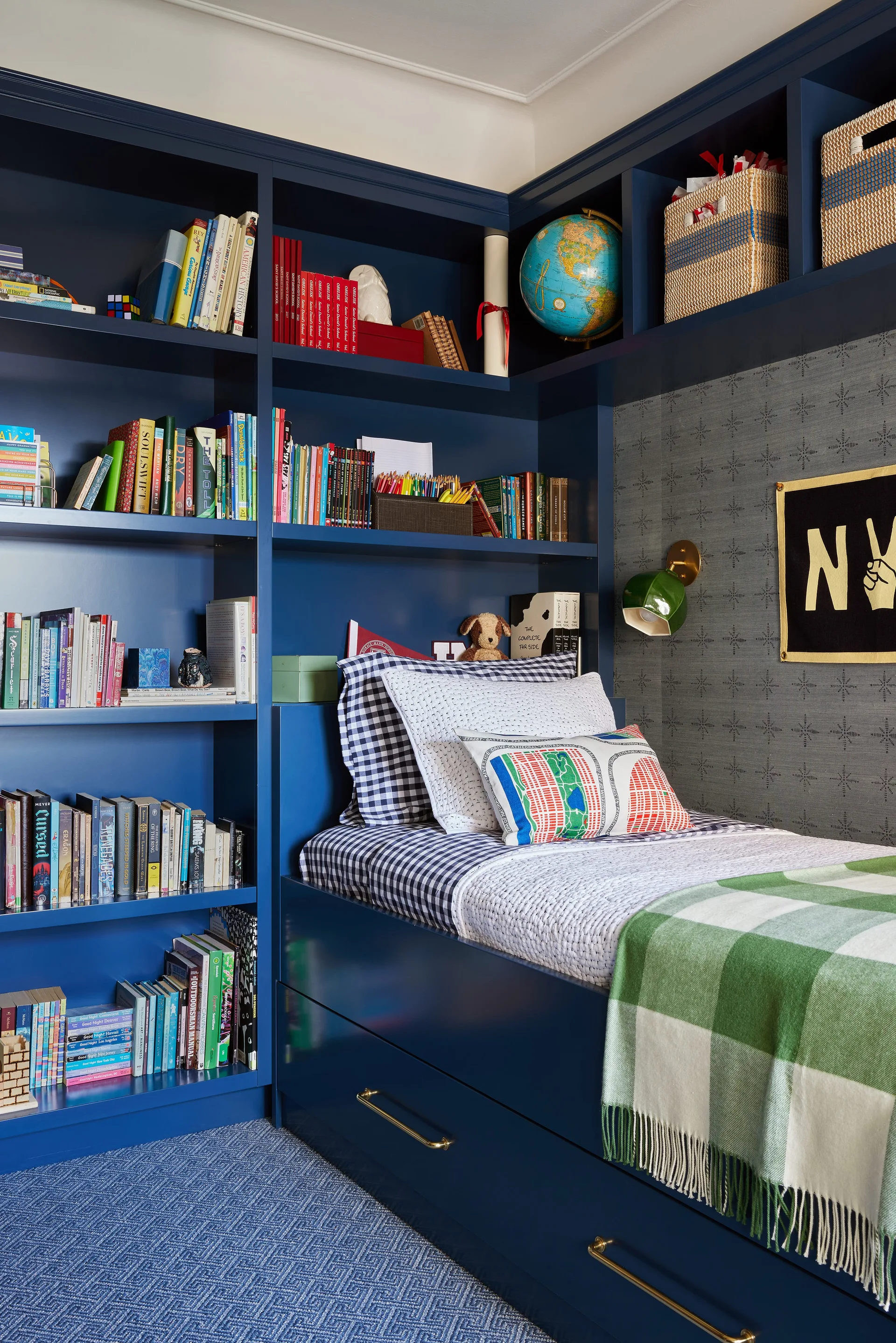 A child's navy blue built-in bed with storage drawers sits next to matching bookshelves filled with books, set against grey patterned wallpaper.