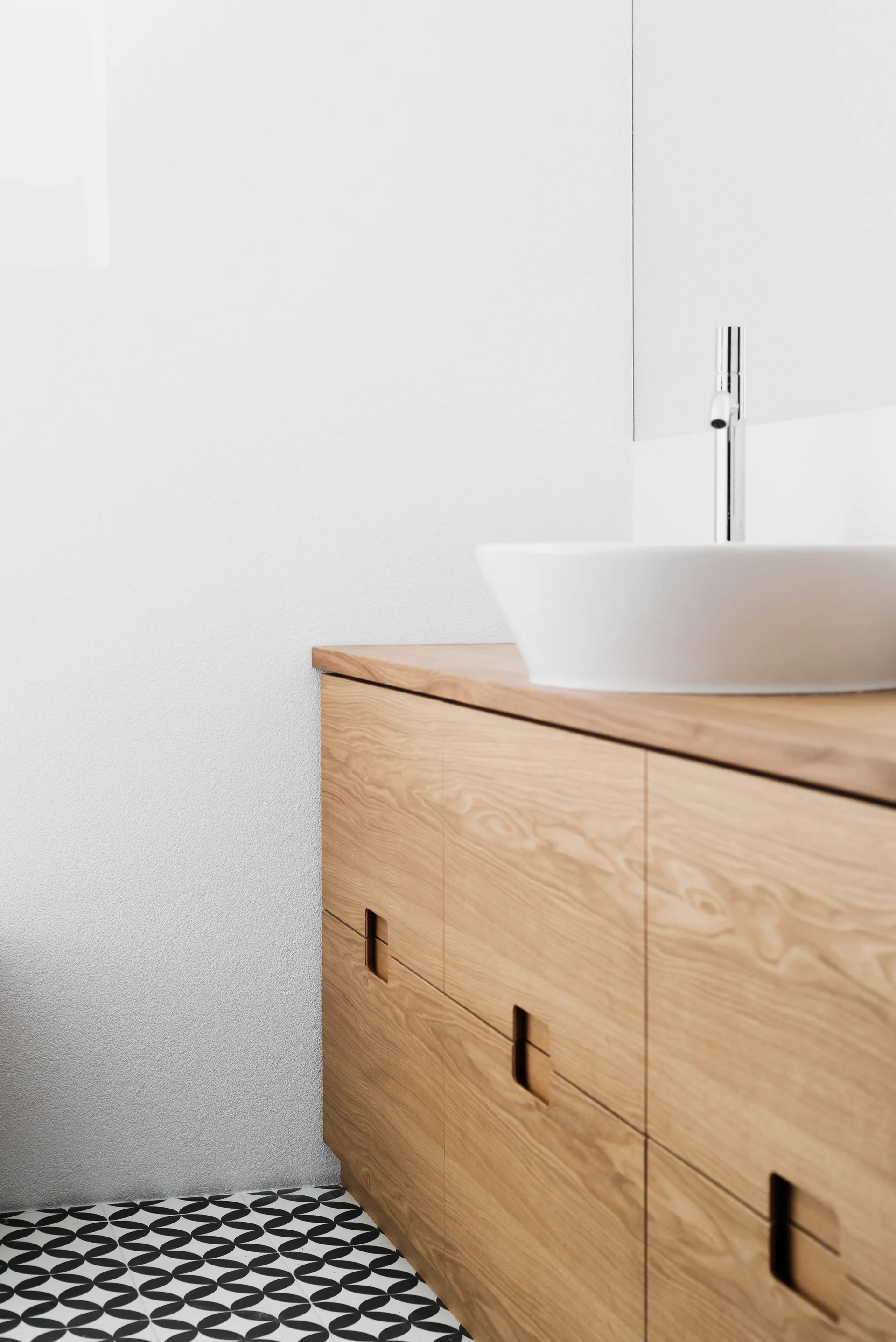 A light wood bathroom vanity with a white vessel sink sits on a black and white patterned tile floor against a plain white wall.