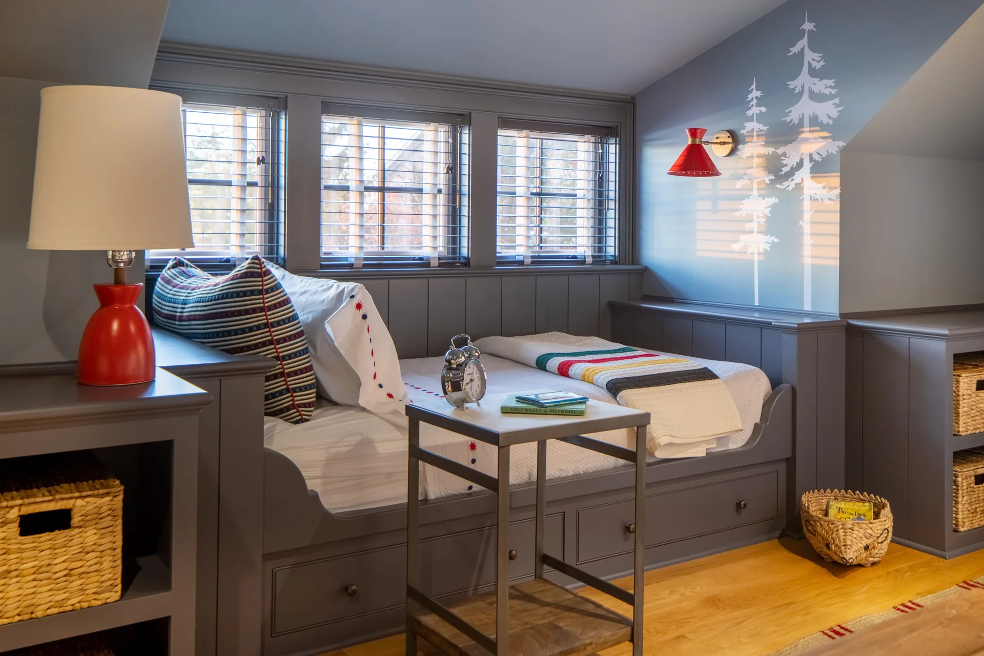 A grey built-in daybed with striped bedding and a red lamp sits beneath three windows with wooden blinds in a cozy bedroom.