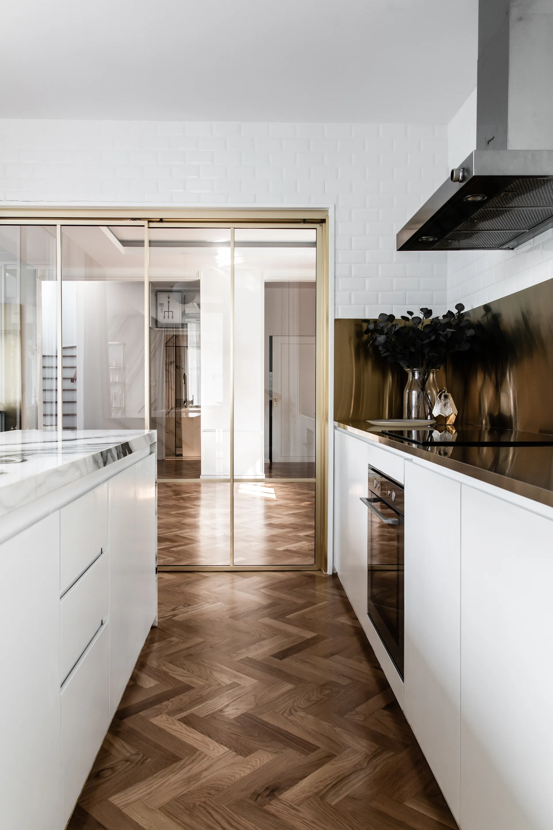A modern kitchen featuring white cabinets, a reflective gold backsplash, a marble-topped island, and a rich wood herringbone floor.