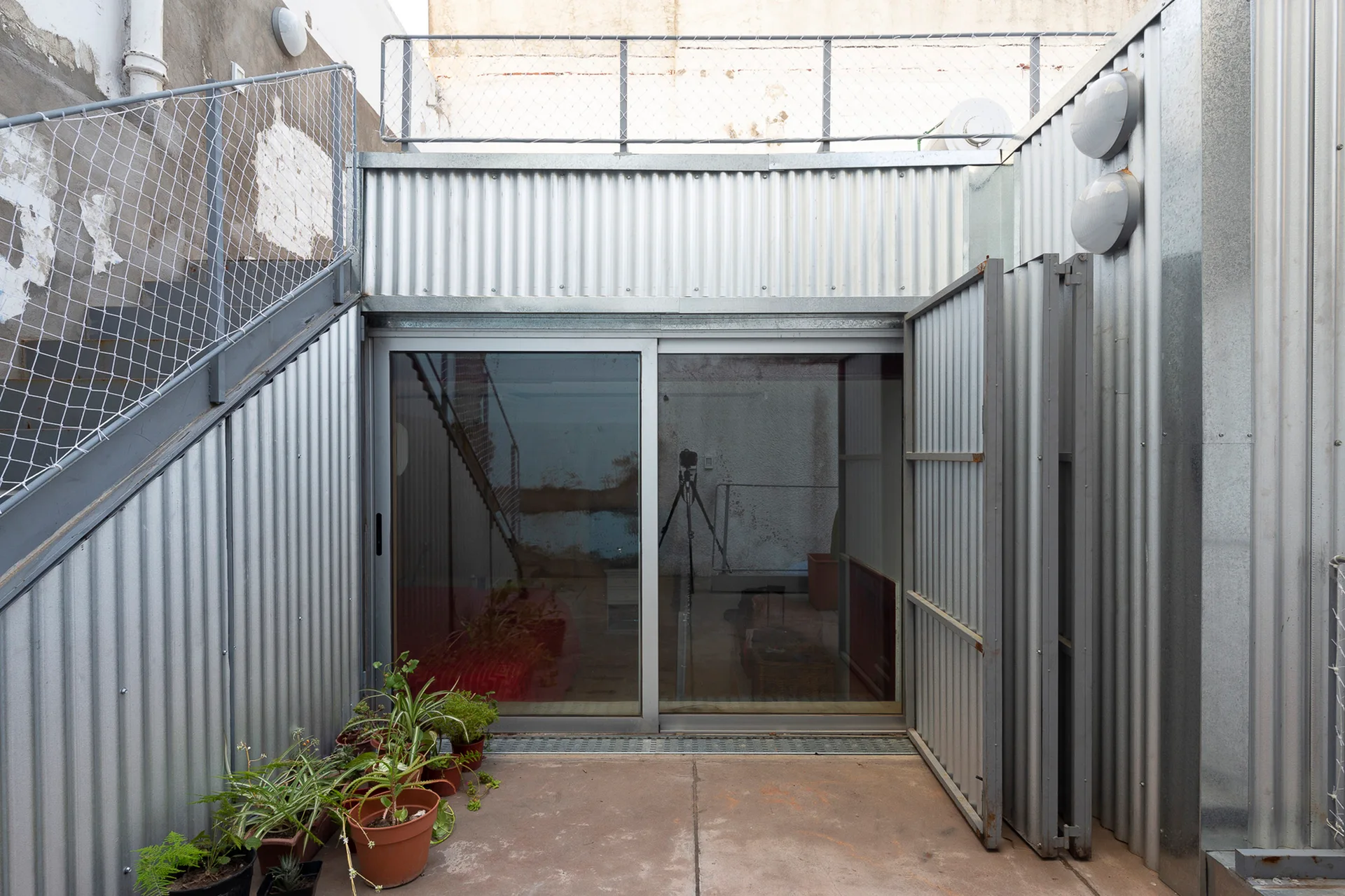 An industrial outdoor patio featuring corrugated metal siding, a sliding glass door, an exterior staircase, and several potted plants on a concrete floor.