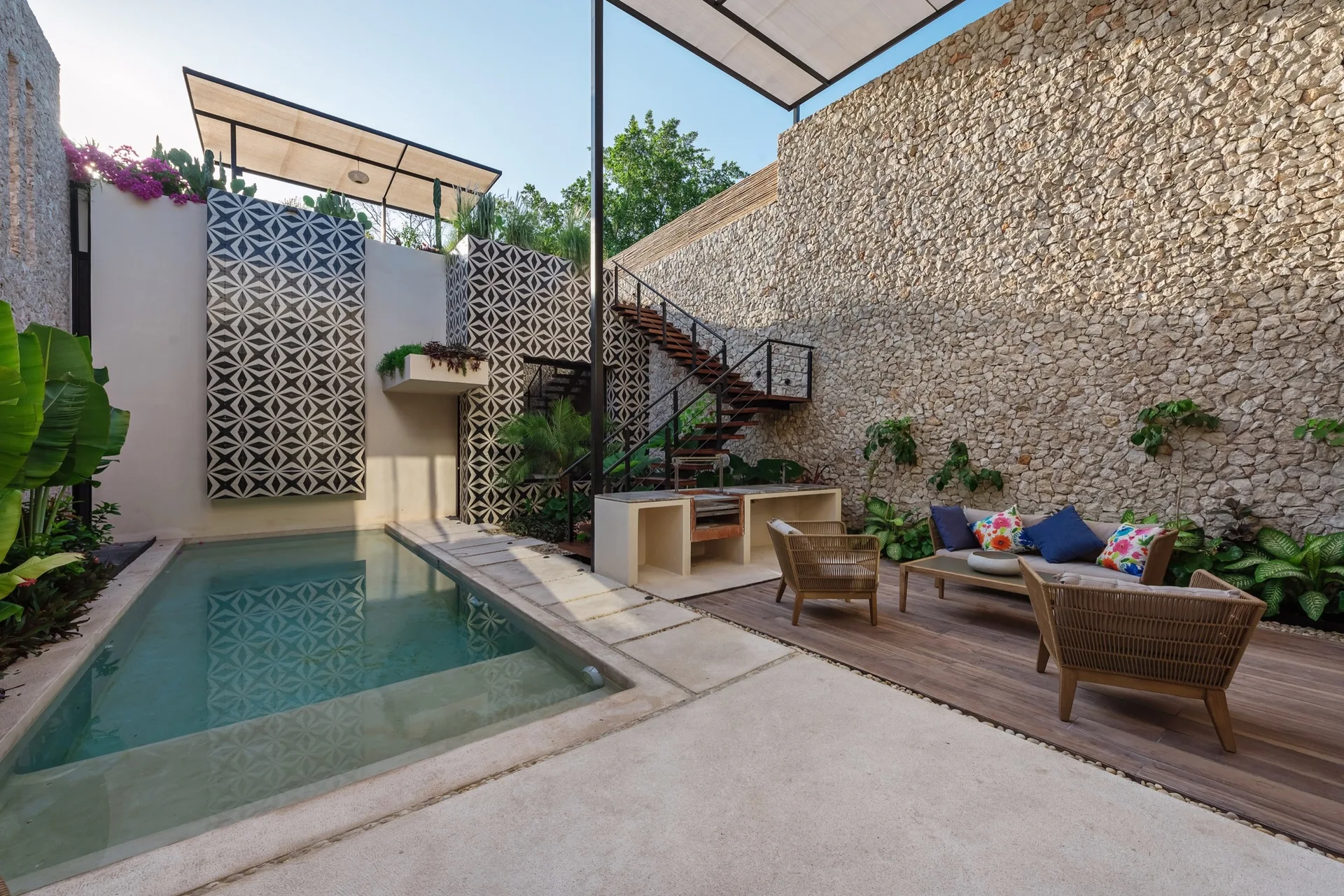 An outdoor patio featuring a rectangular swimming pool next to a wooden deck with wicker seating, set against a tall stacked-stone wall.
