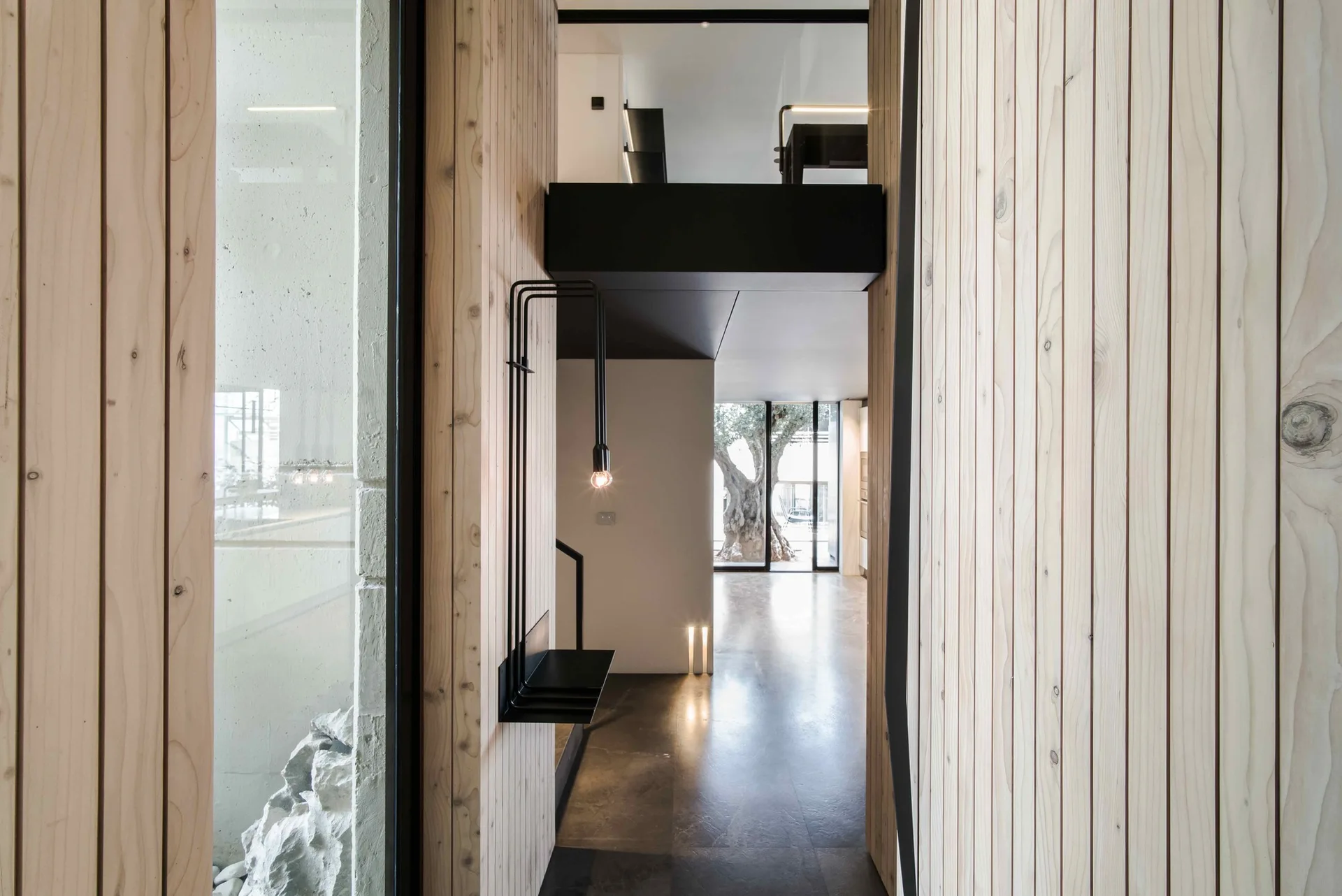 A modern interior corridor features vertical light wood plank walls, a dark concrete floor, and a minimalist black light fixture.