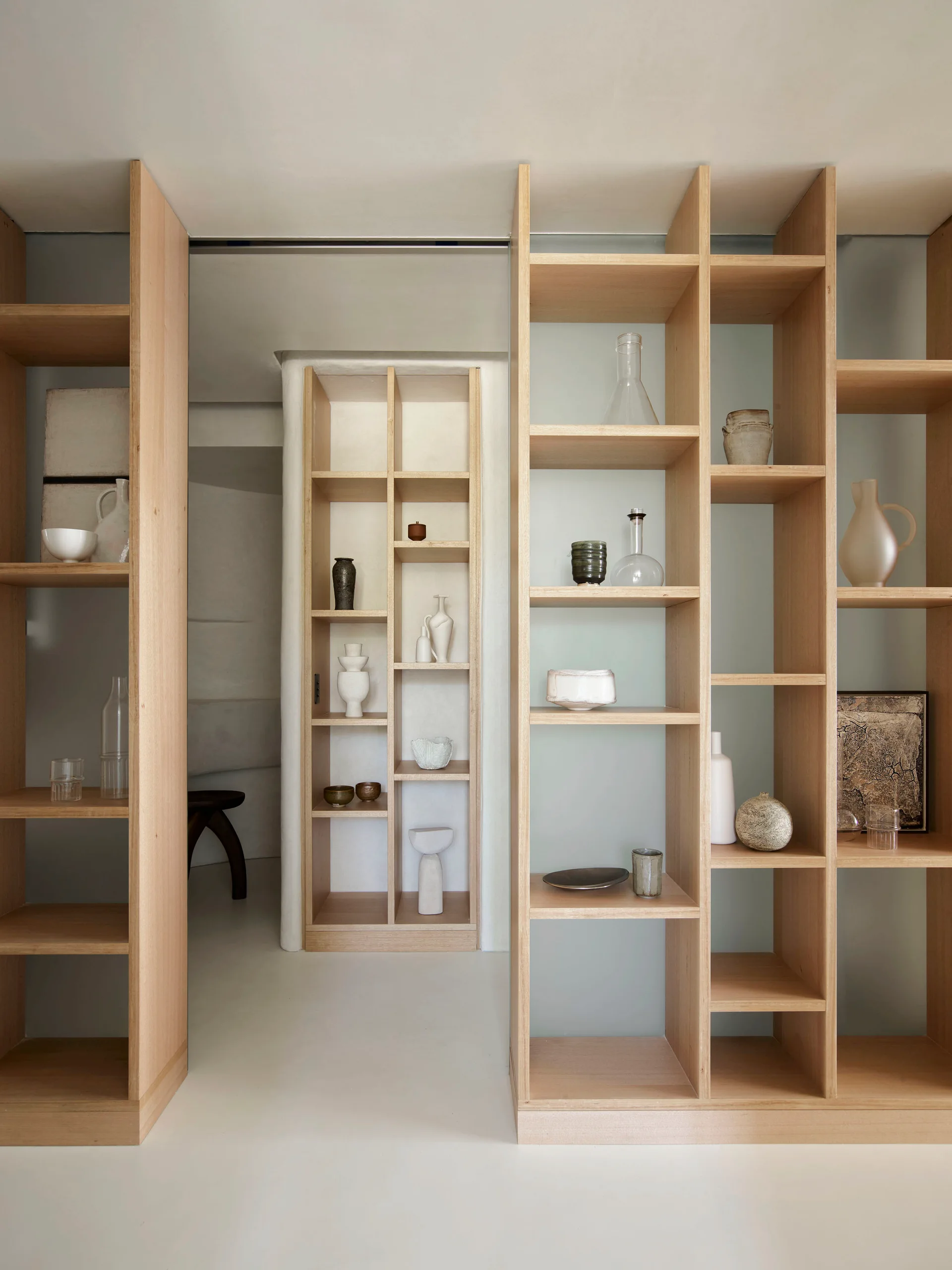 Custom light wood open grid shelving units filled with minimalist ceramic vases and glass vessels act as room dividers against off-white walls.