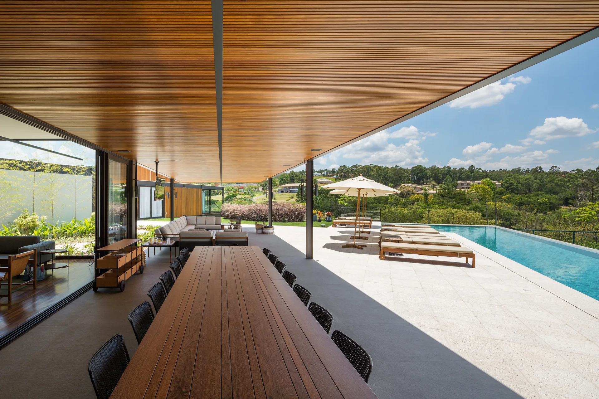 A long wooden dining table and black chairs sit under a slatted wood ceiling on a covered patio next to an infinity pool and lounge chairs.