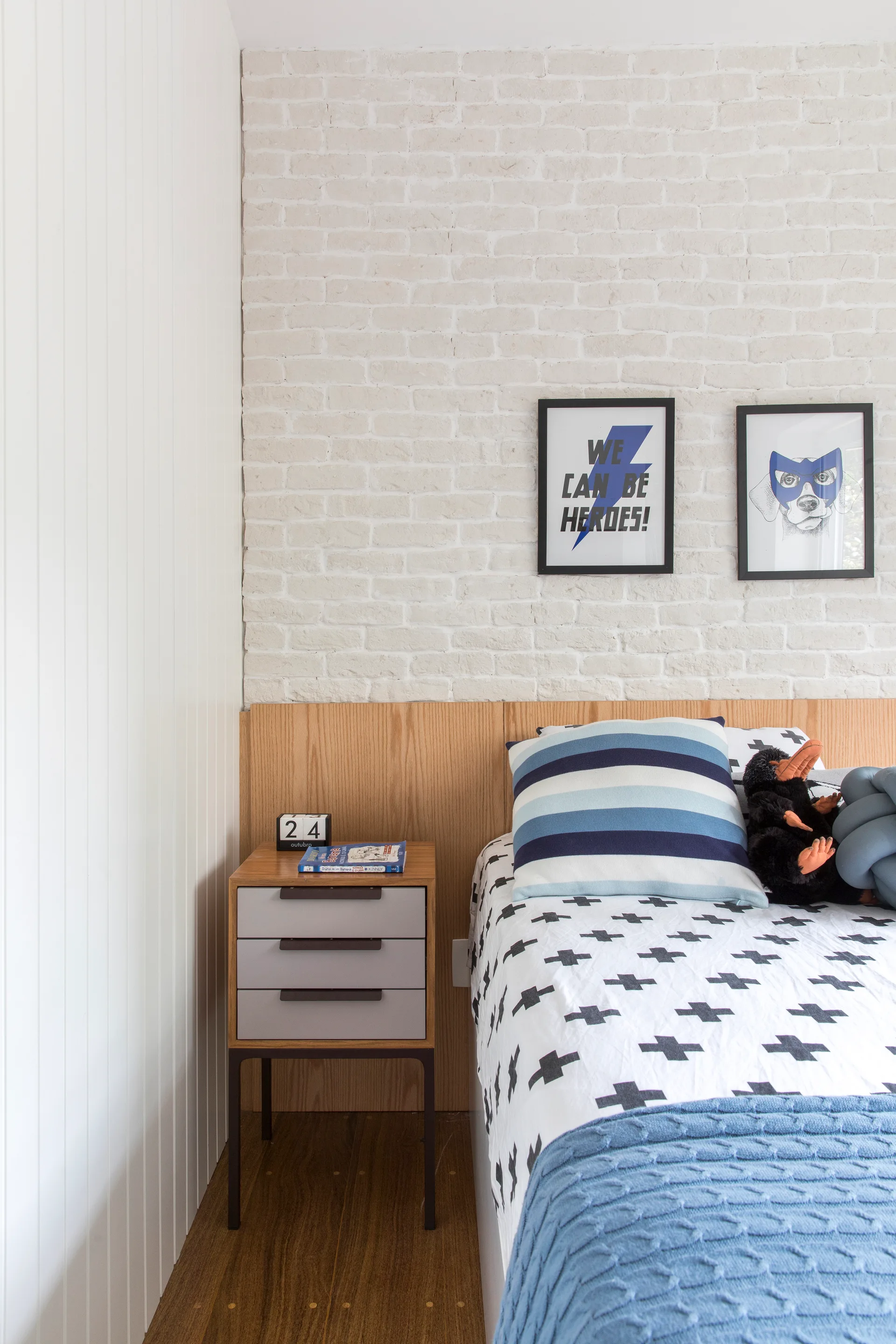 A kids bed with geometric sheets sits against a low wood headboard and a white brick wall featuring two framed graphic art prints.