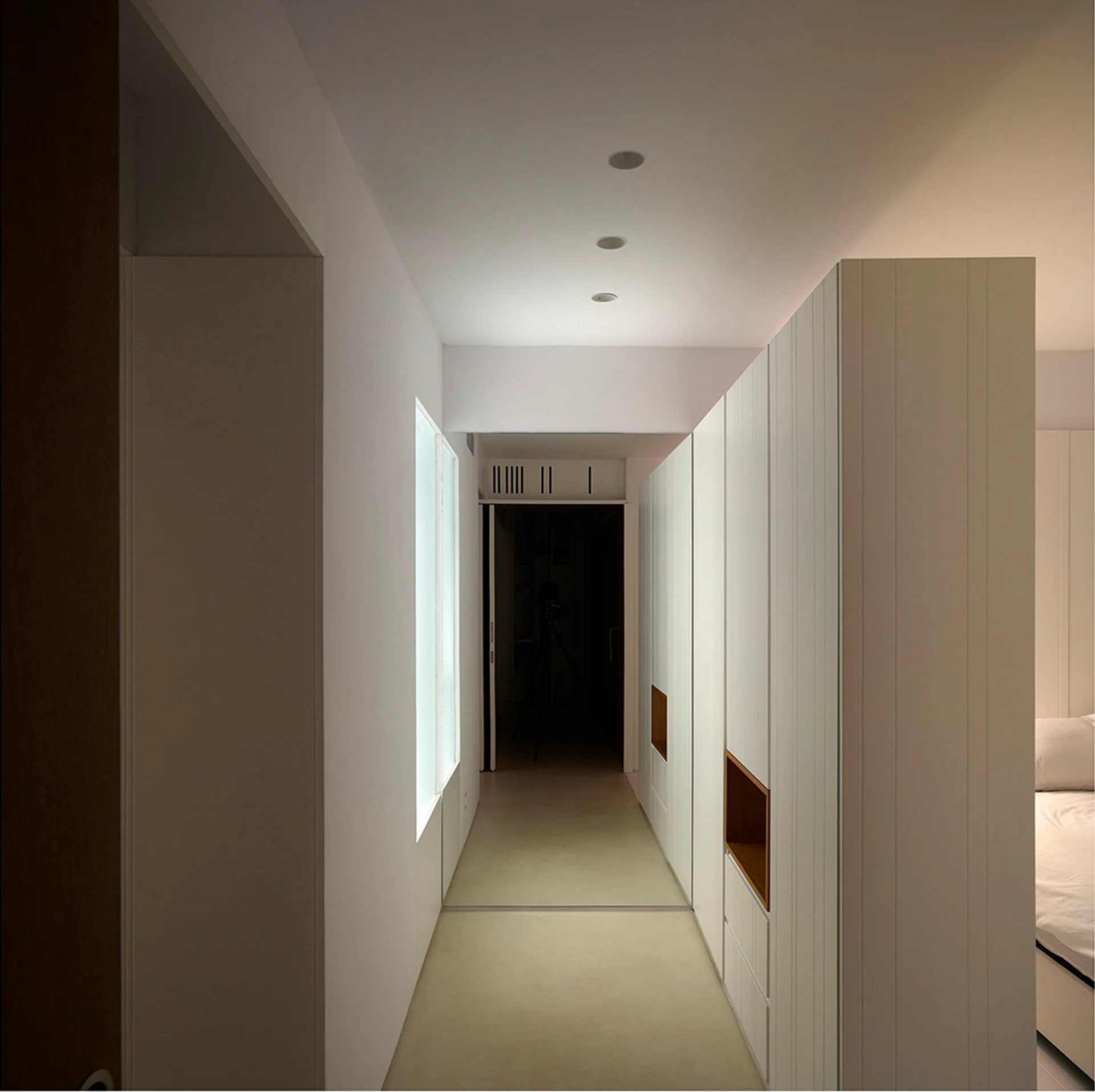 A minimalist white hallway features tall built-in wardrobes with vertical paneling, recessed wooden niches, and a vertical translucent window on the left.