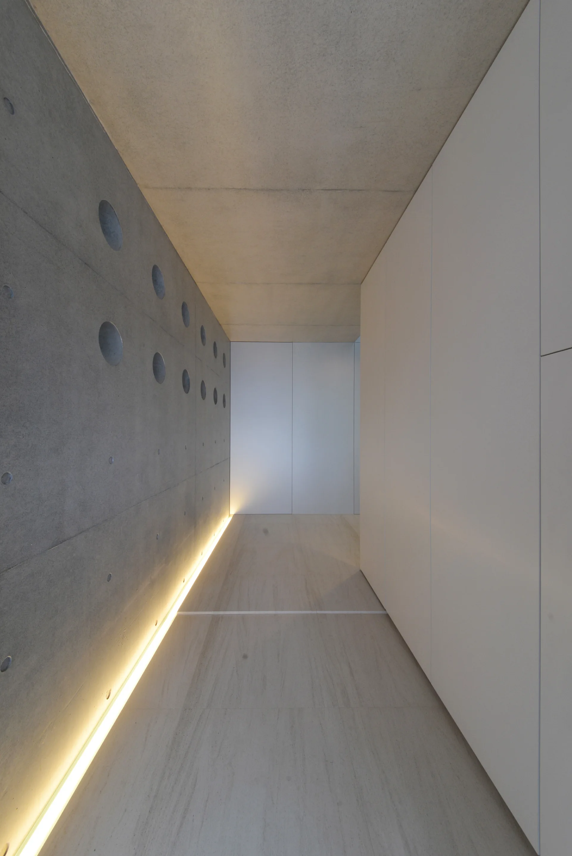 A minimalist corridor featuring a textured concrete wall with circular perforations on one side and seamless white cabinets on the other.