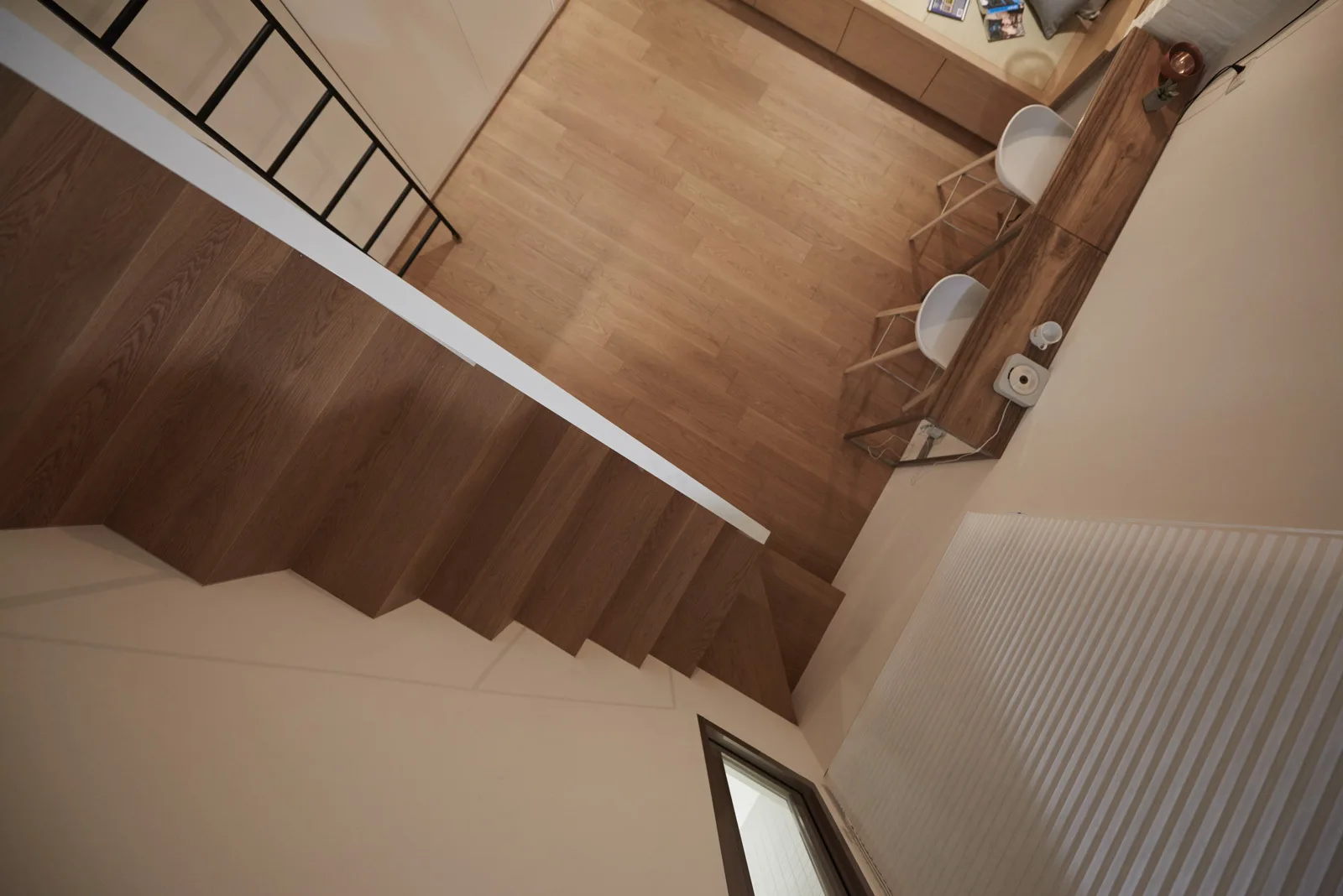High-angle view of a modern loft space with a wooden staircase, matching wood floors, a slim wood console table, and two white stools.