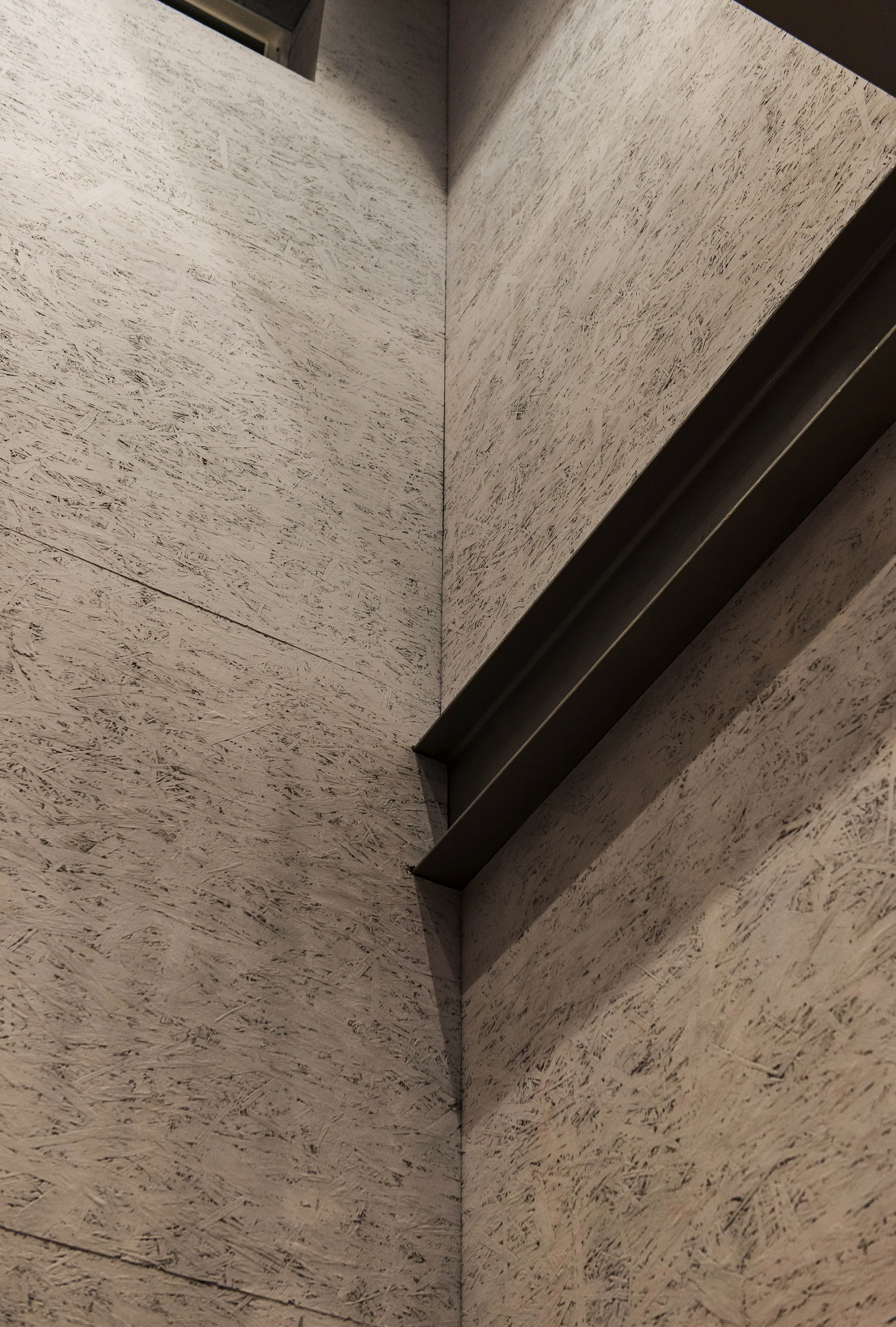 An interior corner with walls made of textured oriented strand board intersected by a horizontal dark grey steel beam under ambient light.