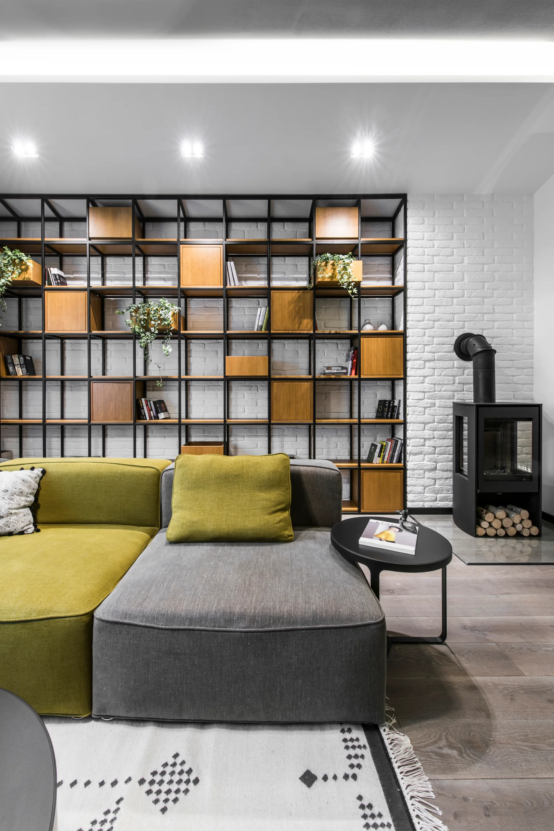 A modern living room features an olive and grey modular sectional sofa, a floor-to-ceiling metal shelving unit, and a black wood-burning stove.
