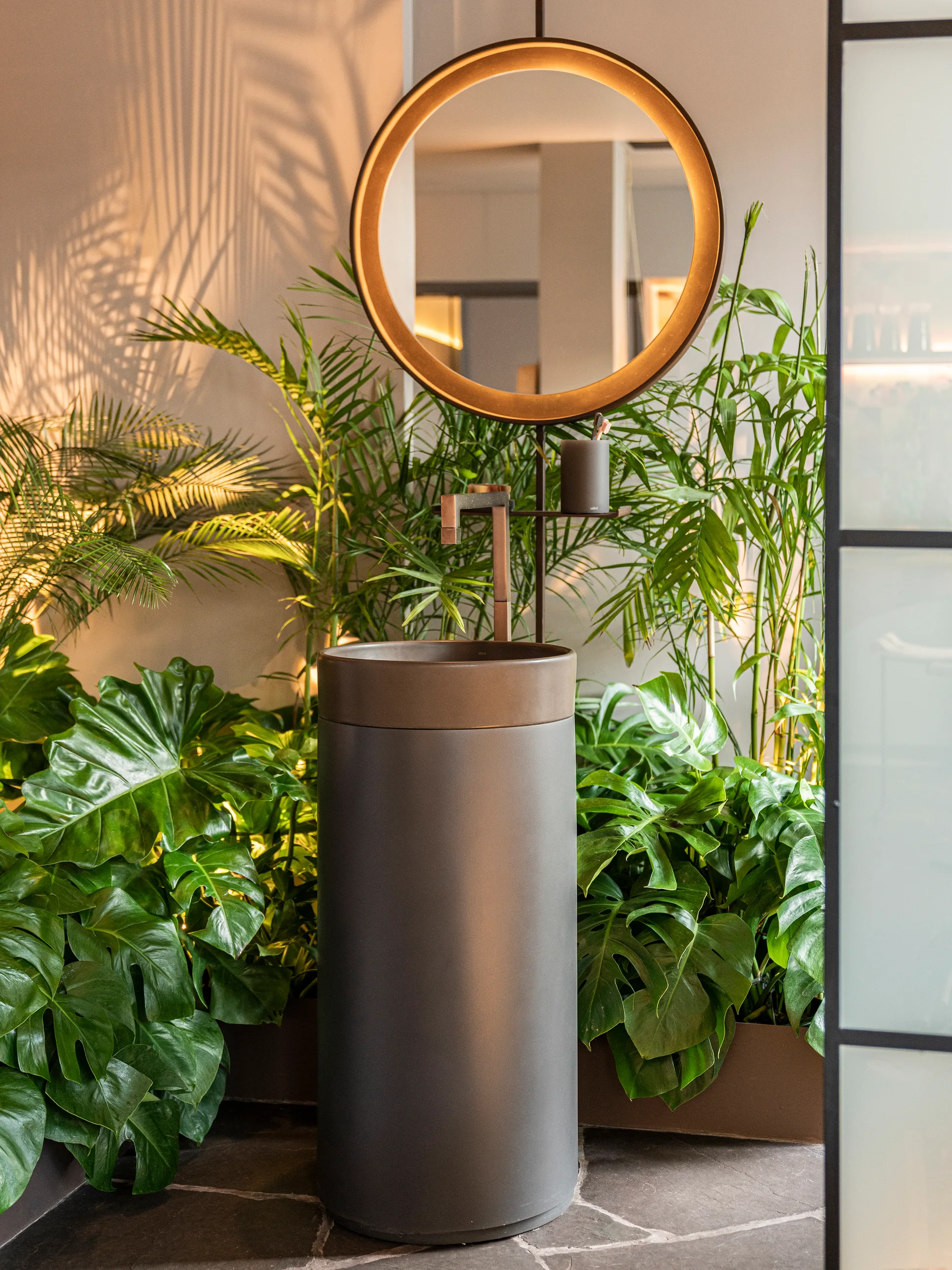 A dark grey cylindrical pedestal sink stands on a flagstone floor surrounded by lush green houseplants under a glowing, backlit round mirror.