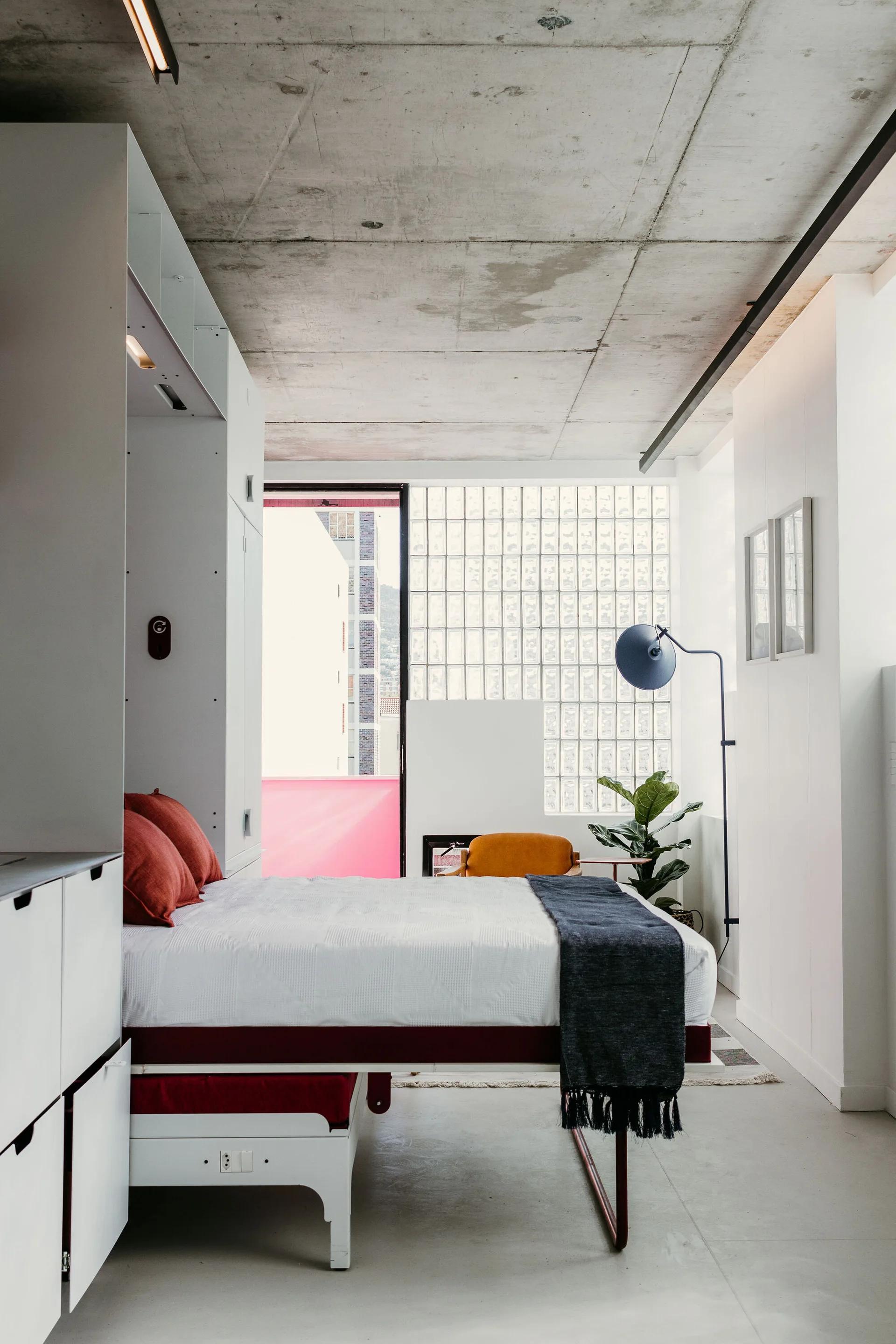 A modern bedroom featuring an integrated white bed and storage unit under a raw concrete ceiling with a translucent glass block wall in the background.