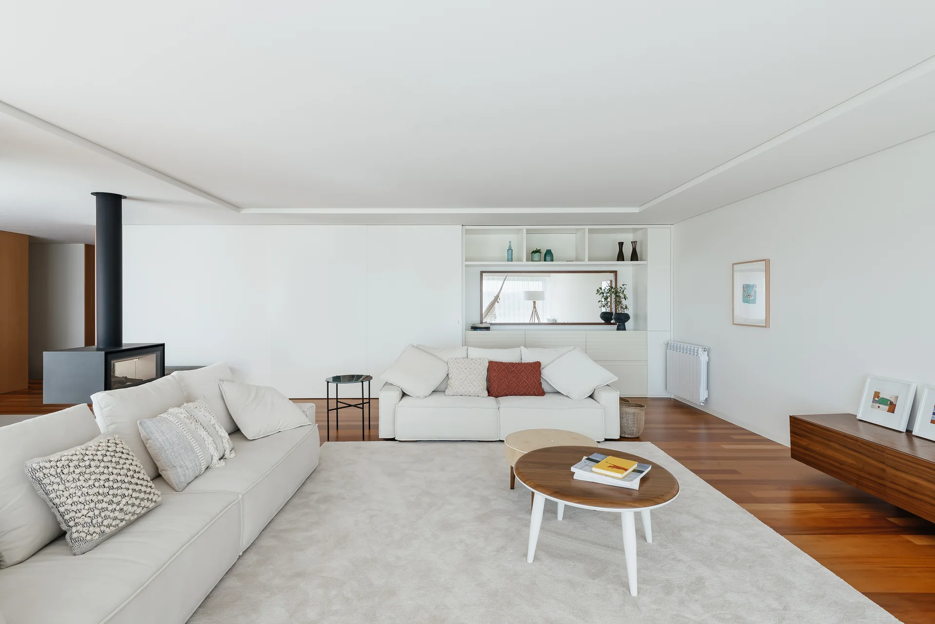 A minimalist living room with white sofas, a large neutral rug over hardwood floors, a black wood-burning stove, and a floating wood console.