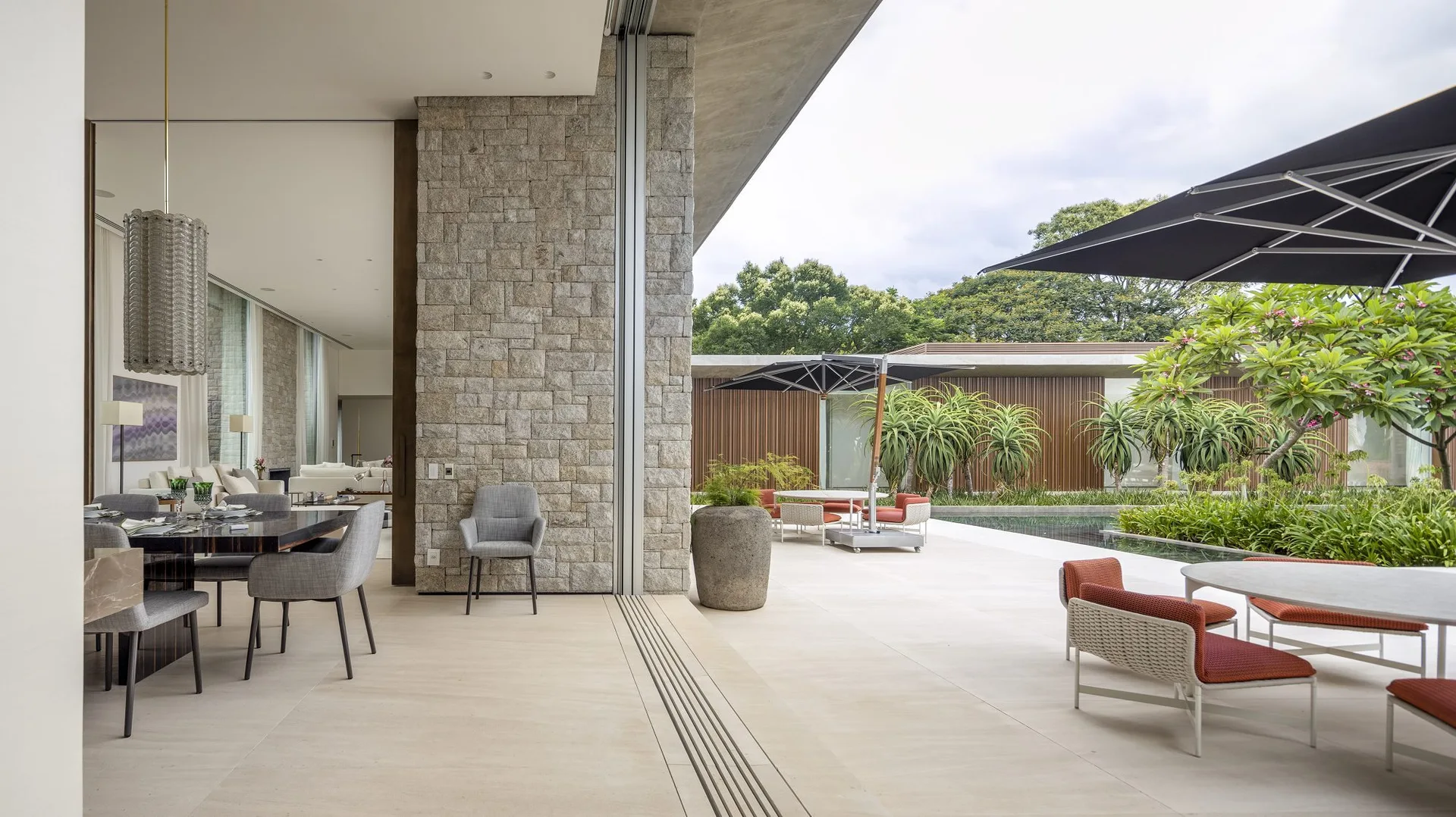 A modern indoor dining area with grey chairs opens up to a spacious outdoor patio with white and orange furniture next to a pool.