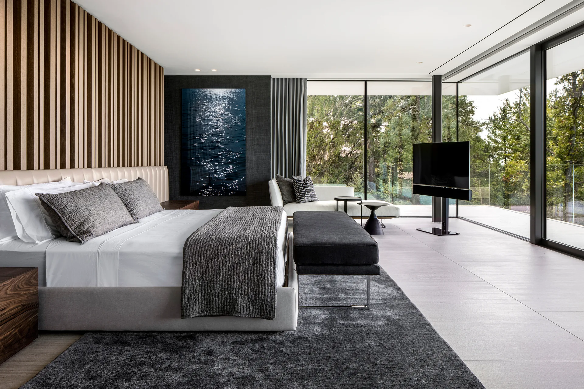 A modern bedroom featuring a wood-slat accent wall, a grey bed with a dark bench, a chaise by floor-to-ceiling glass walls, and forest views outside.
