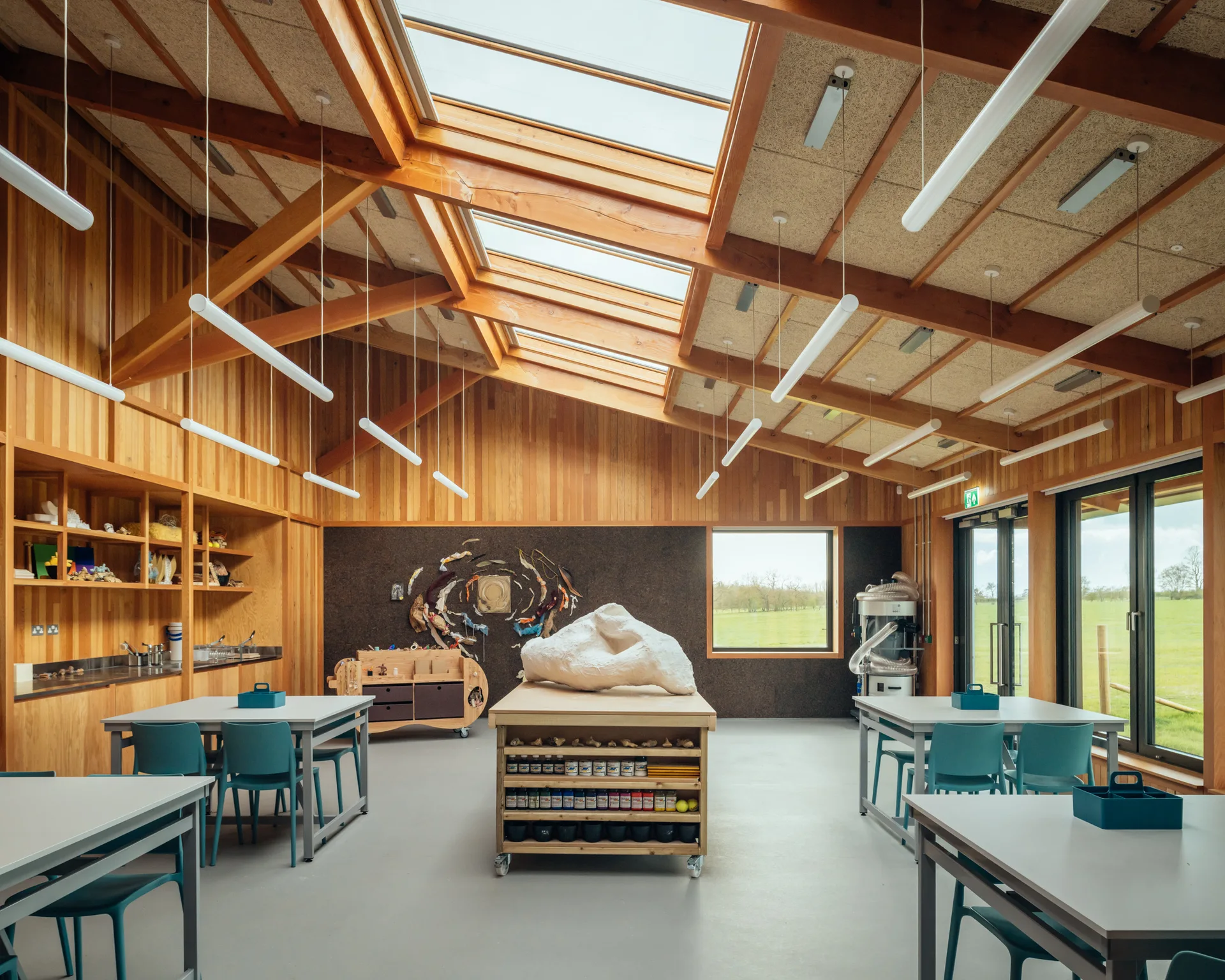 A spacious art studio features natural wood-plank walls, exposed timber ceiling beams, large skylights, and multiple worktables with teal chairs.