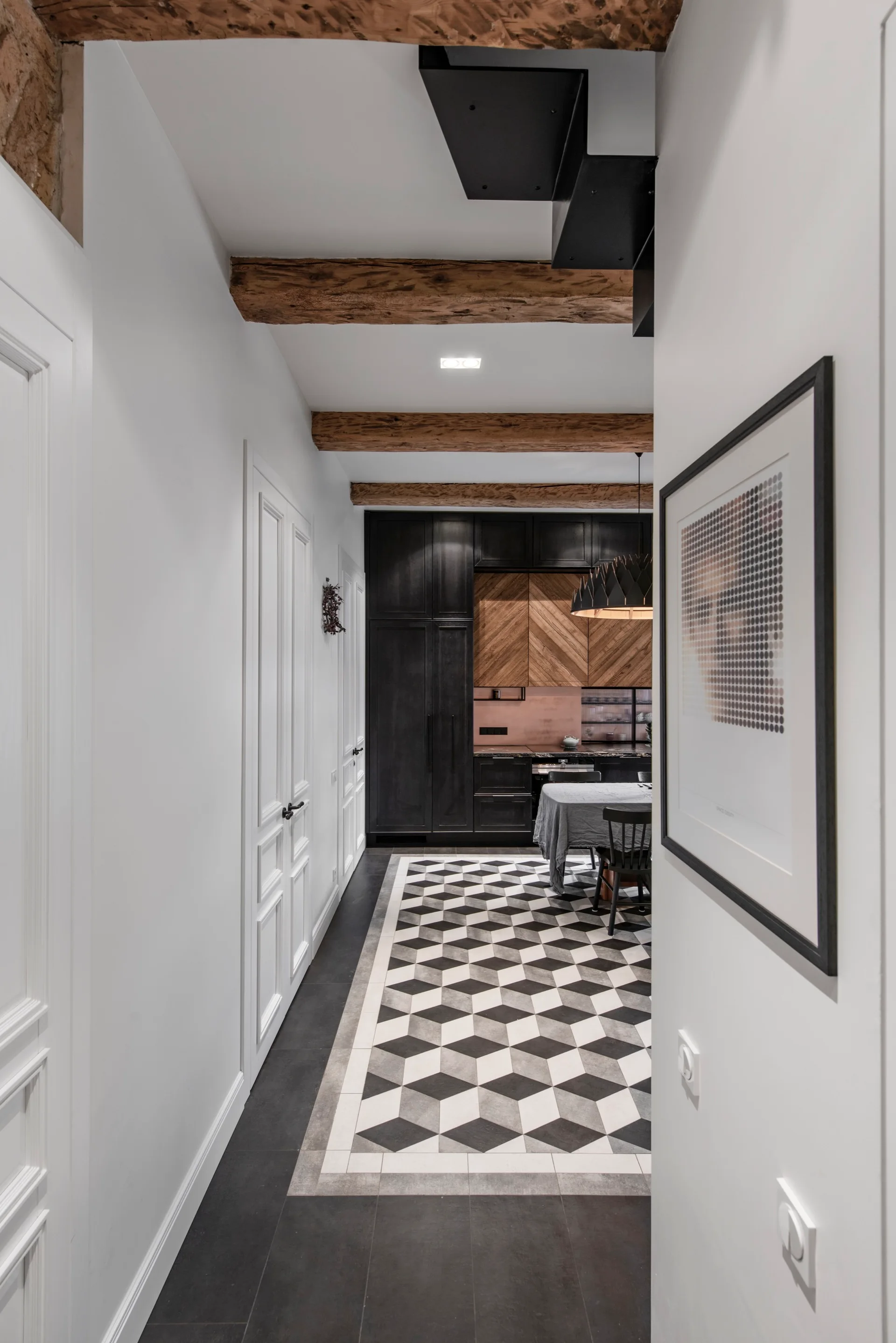 A view through a white hallway leads to a kitchen featuring geometric cube floor tiles, matte black cabinets, and rustic wood ceiling beams.