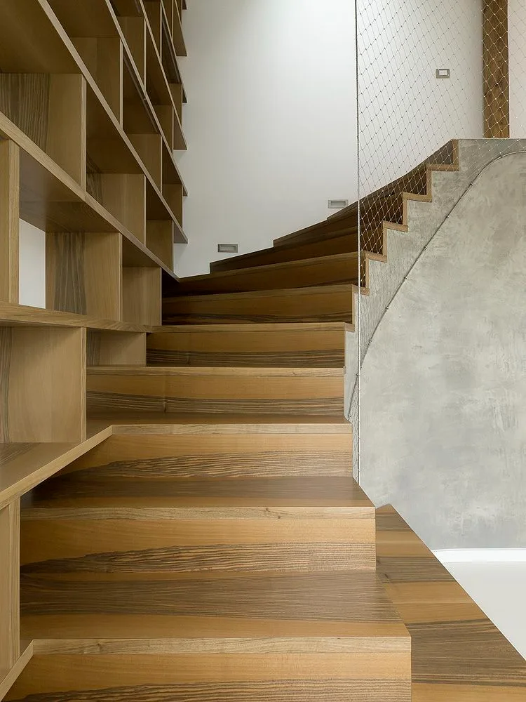 A winding wooden staircase with integrated shelving on one side and a textured concrete wall with a metal wire mesh guard rail on the other.
