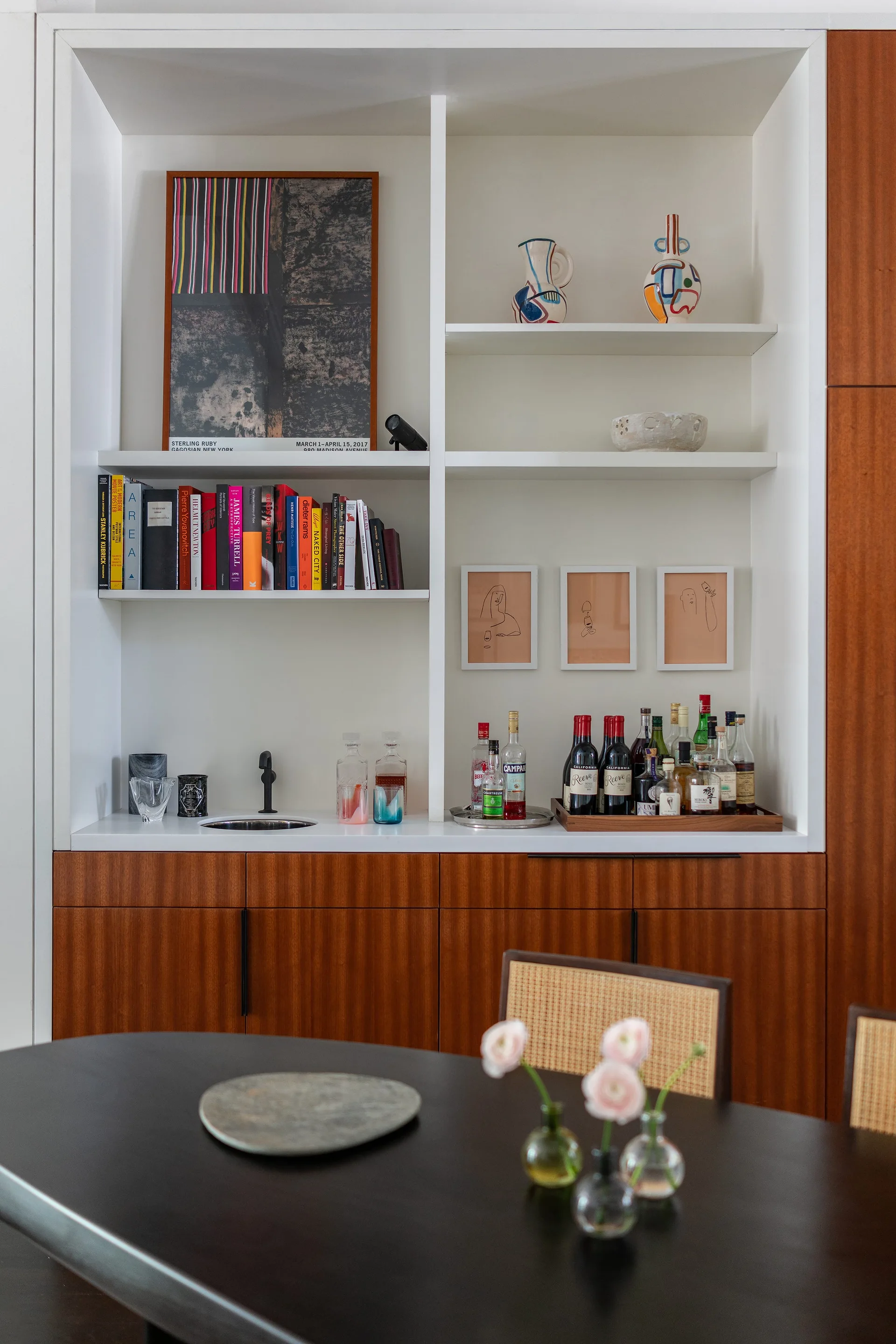 A built-in home bar features warm wood cabinets, white open shelves with books and art, and a small black sink on the counter.
