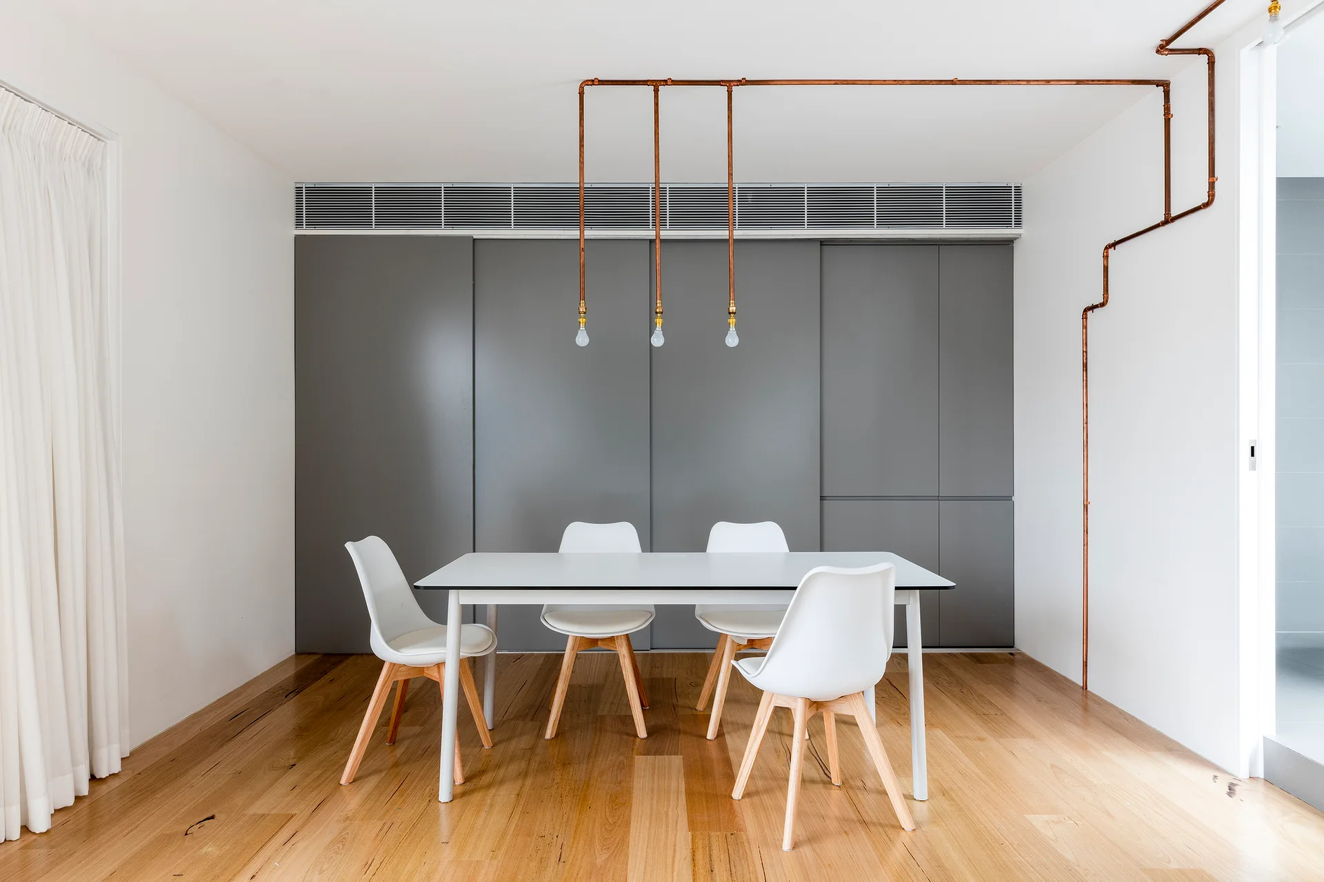 A white dining table and four chairs stand before a grey sliding panel wall, accented by exposed copper pipe lighting from the ceiling.