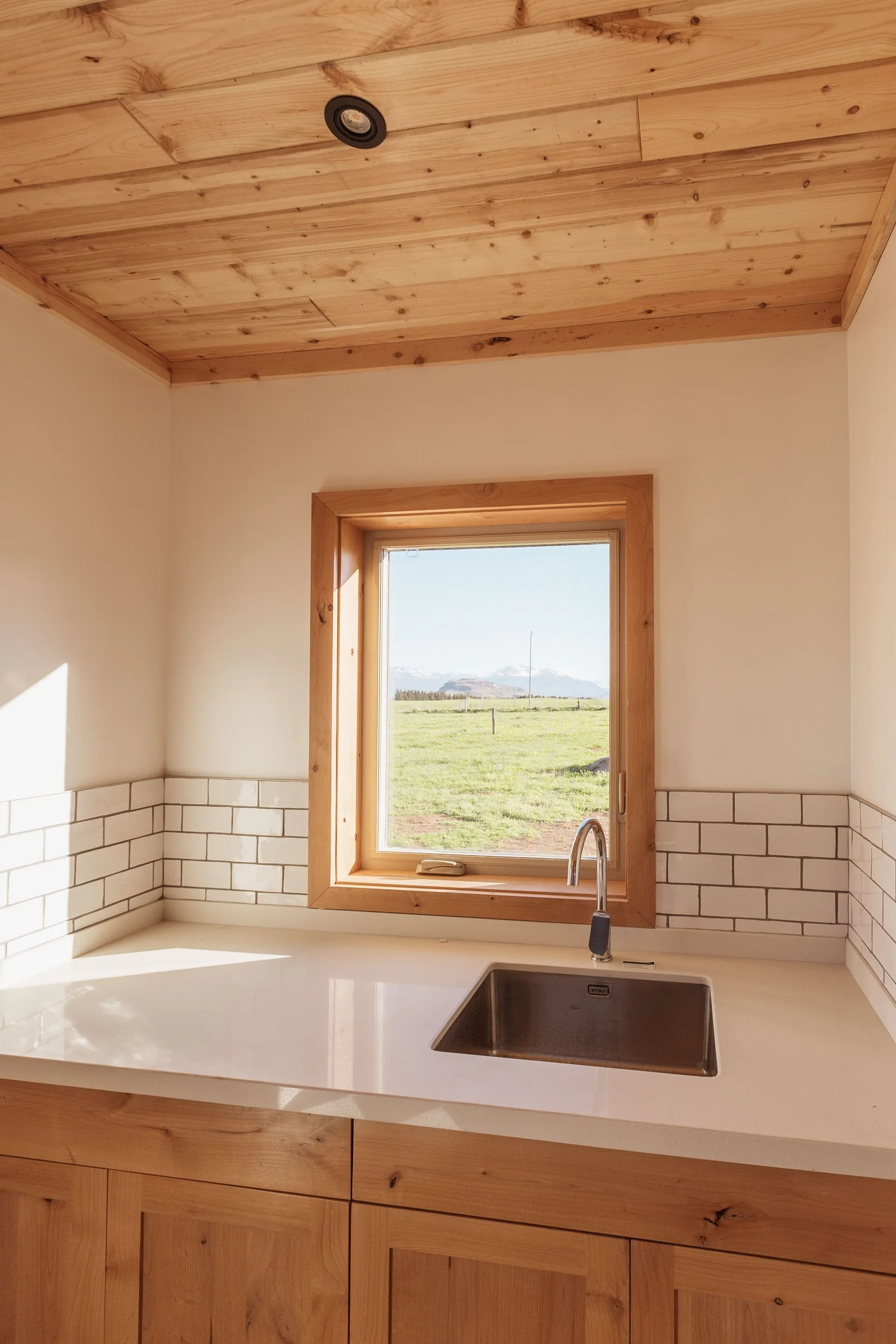 A kitchen with light wood cabinetry, a plank ceiling, a white countertop with an undermount sink, and a window overlooking a green field.