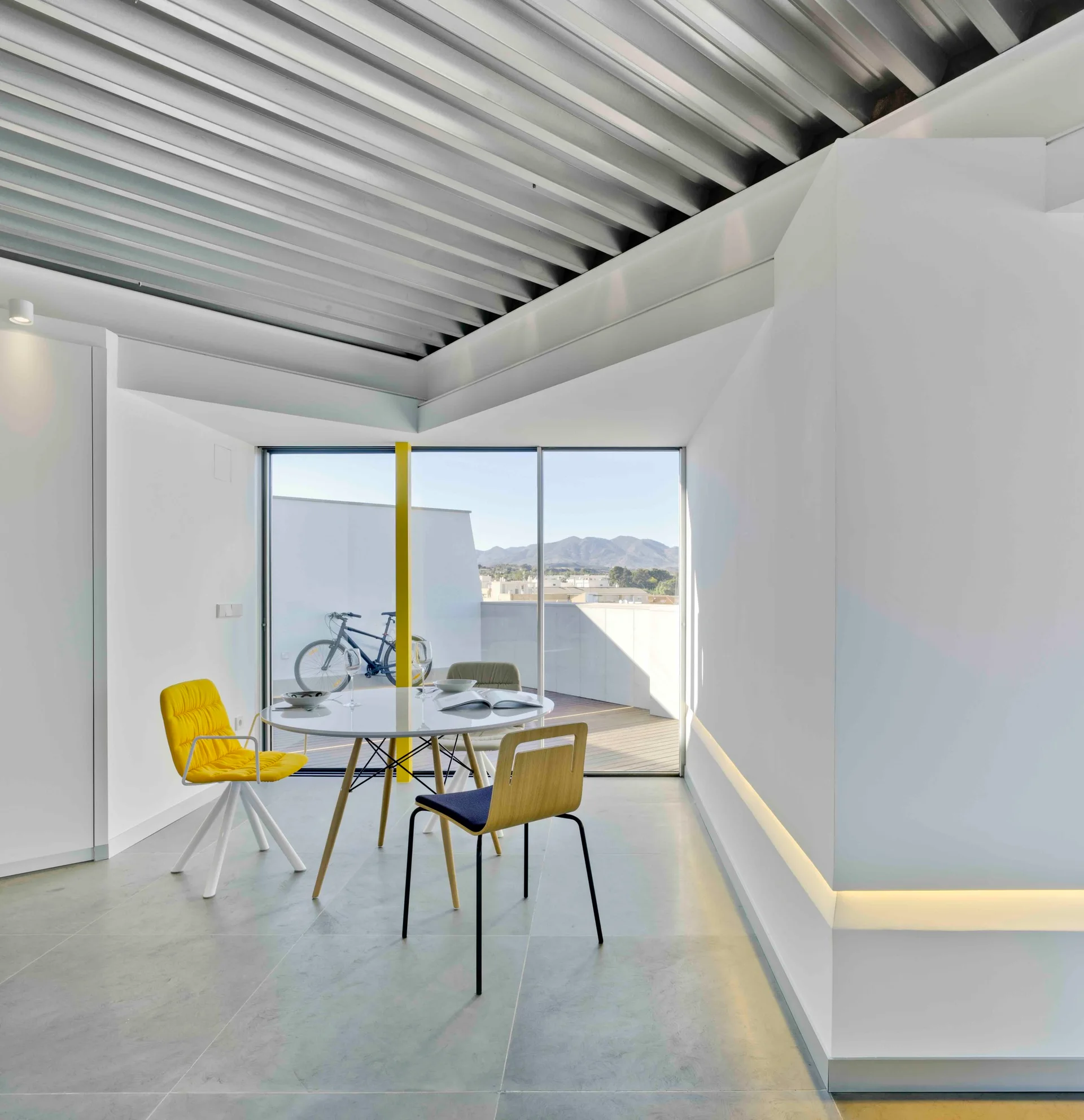 A modern minimalist dining area with a round white table, a yellow chair, and a corrugated metal ceiling next to terrace doors.