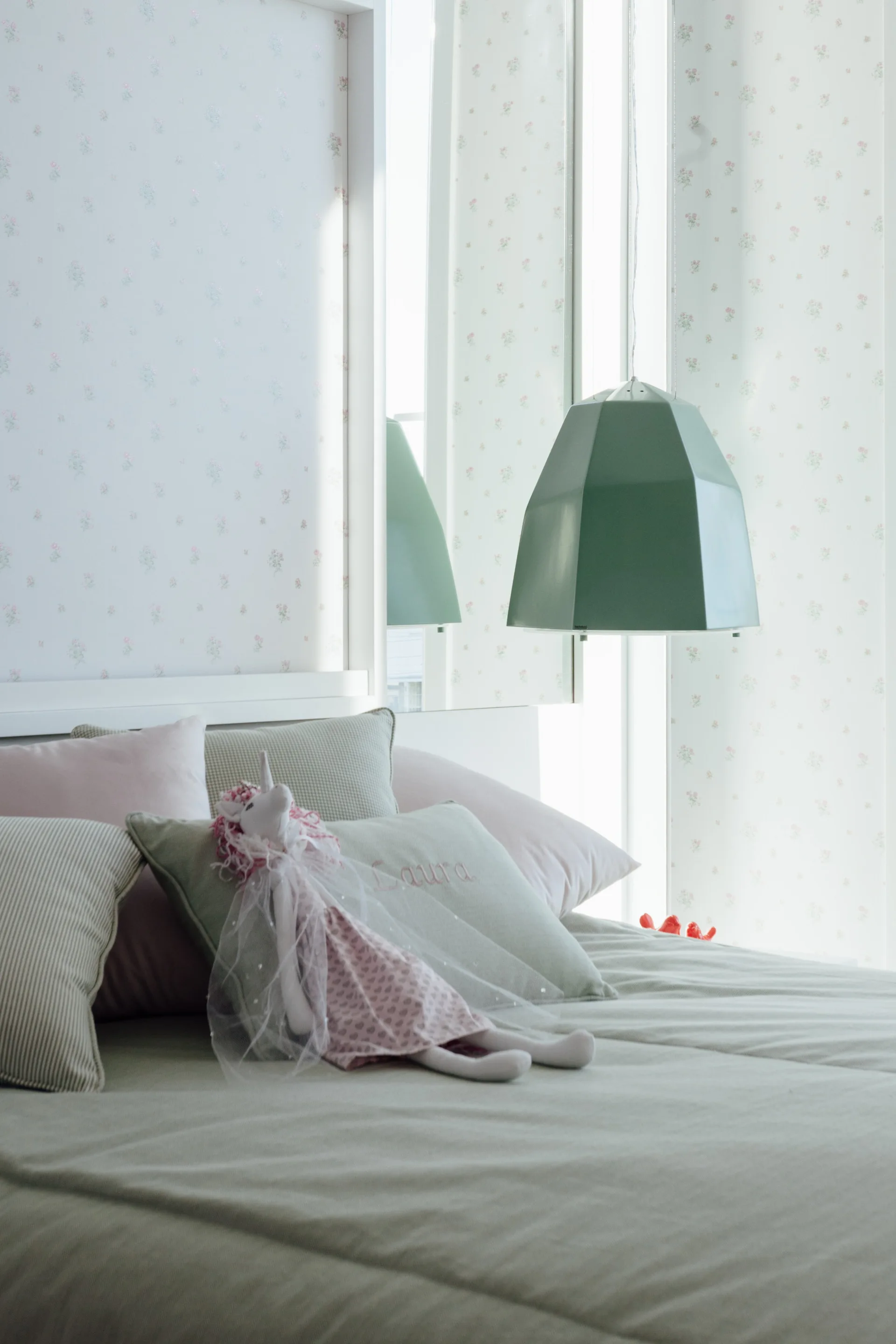 A children's bed with pastel green and pink pillows sits in front of delicate floral wallpaper, illuminated by a geometric green pendant light.