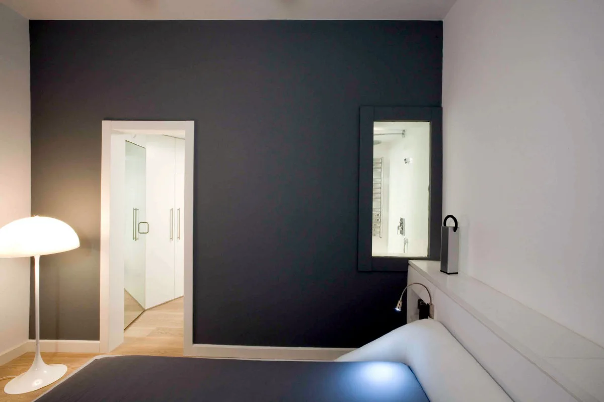 A minimalist bedroom featuring a dark charcoal wall, a white built-in headboard shelf, and an interior window viewing into a brightly lit bathroom.