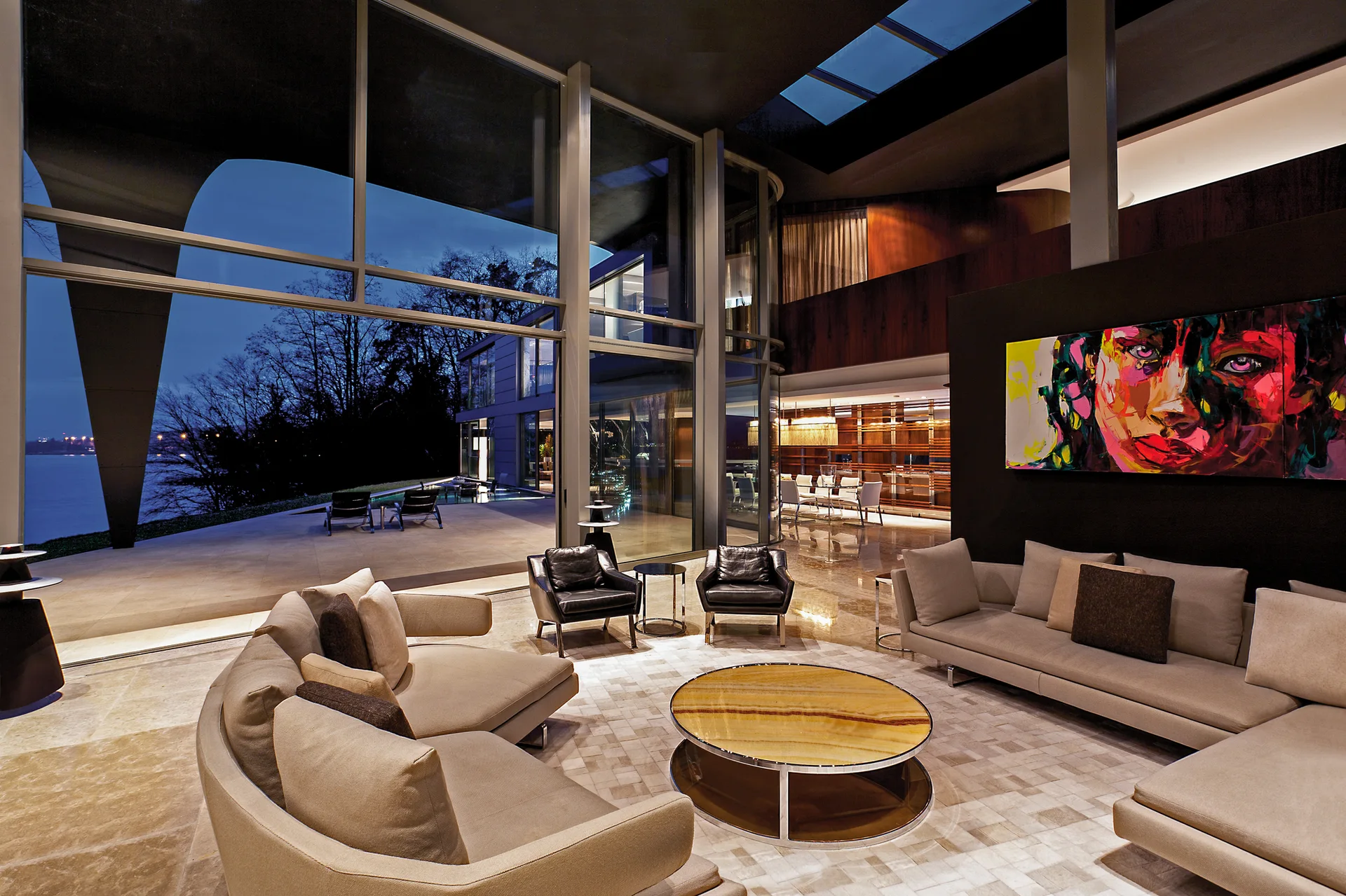 A modern living room features curved modular beige sofas, a round onyx-topped coffee table, and massive glass walls overlooking a lake at dusk.