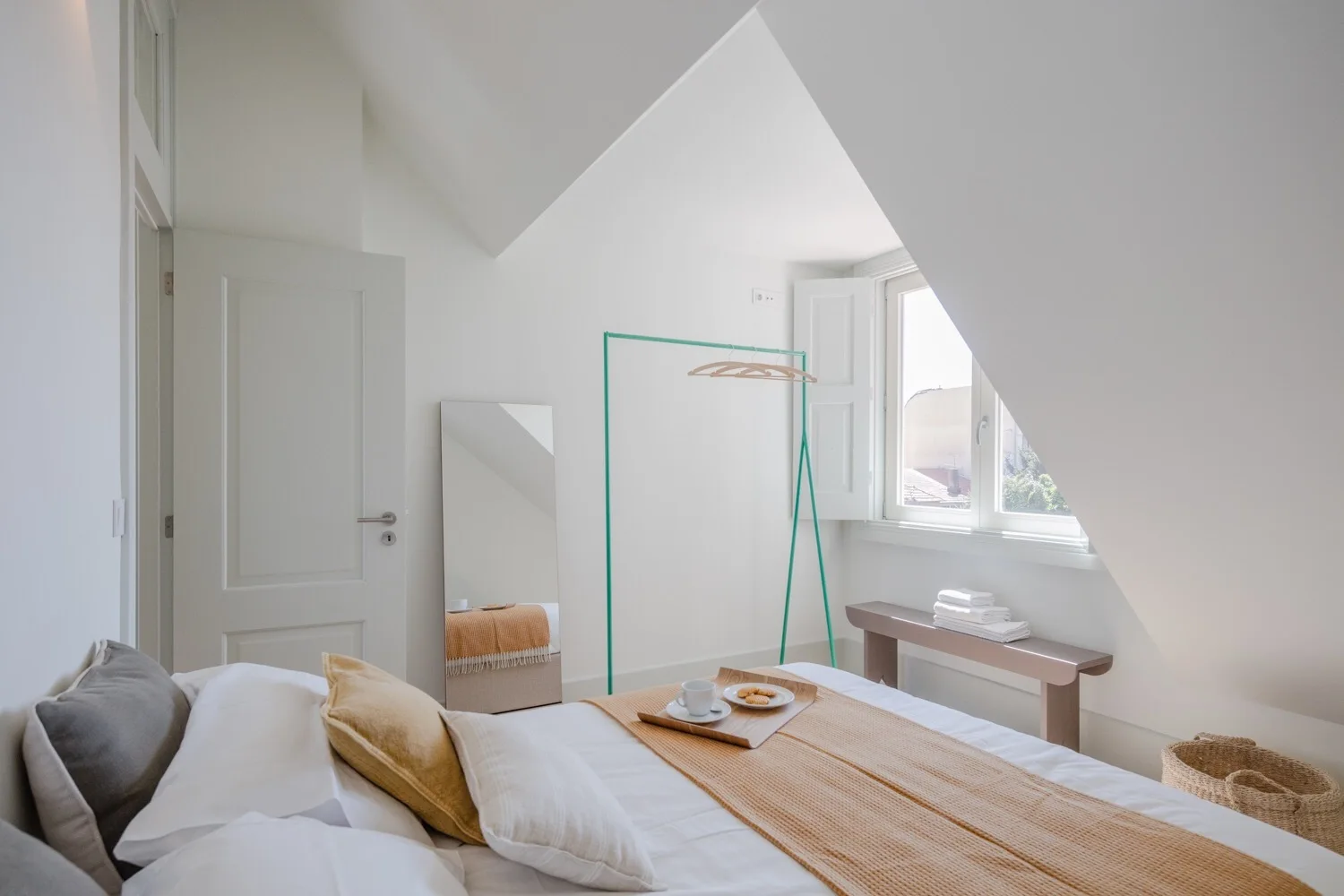 A minimalist attic bedroom features a bed with ochre bedding, a standing mirror, a teal clothing rack, and a bench under the window.