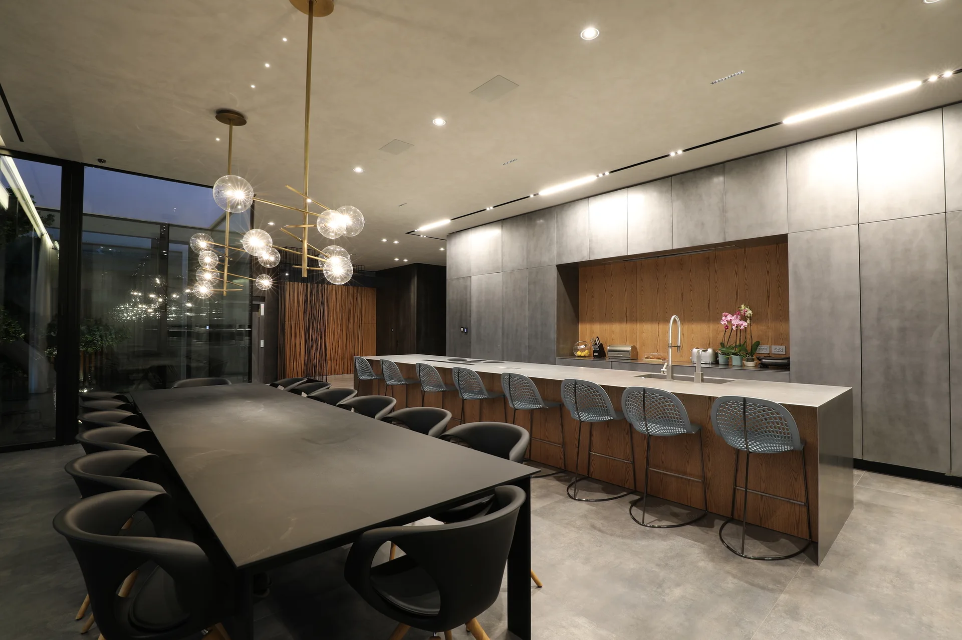 A modern grey kitchen features flat-panel cabinets with a wood-lined alcove, a long island with bar stools, and a large black dining table.