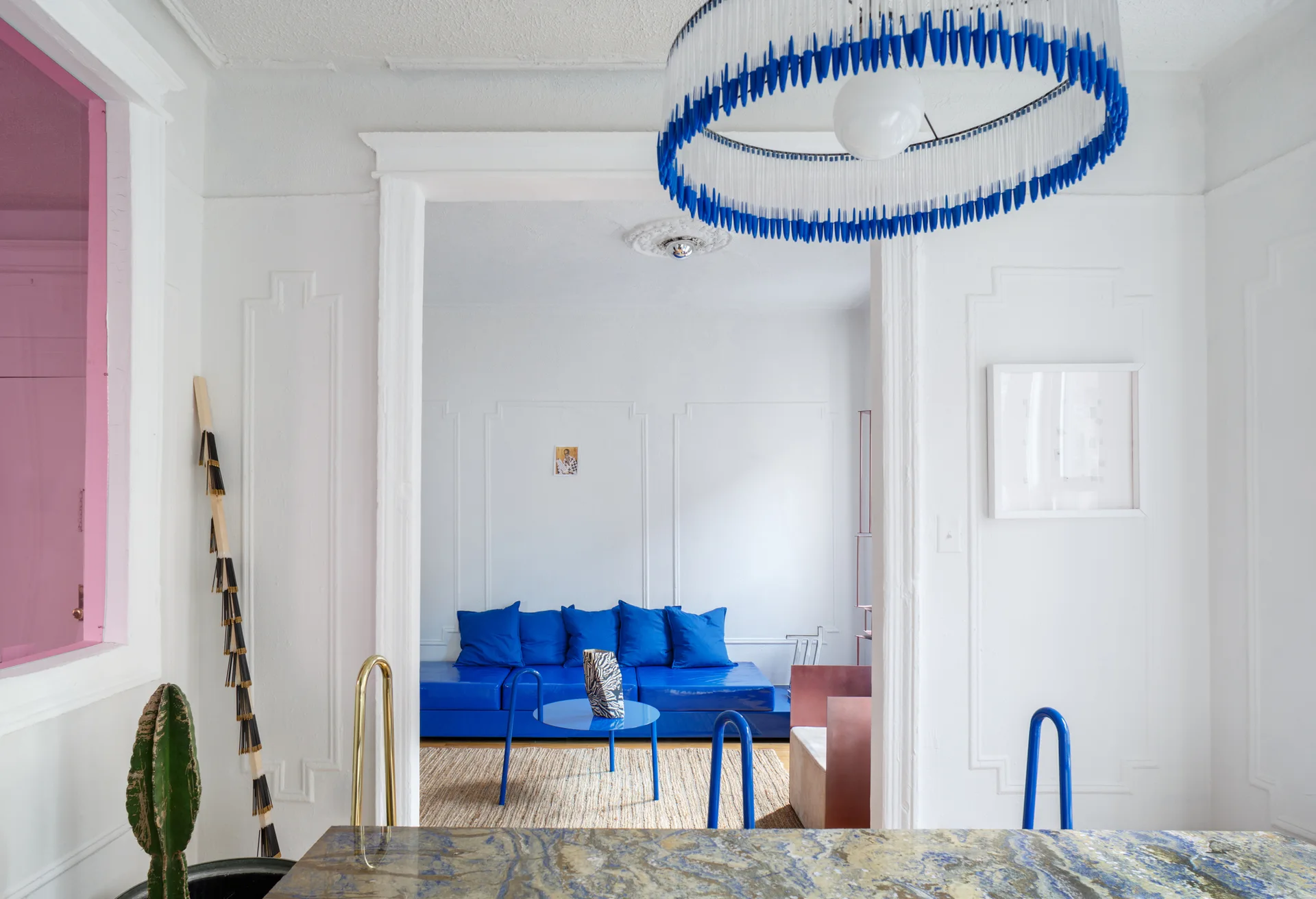 A white living room viewed through an archway, featuring a bright blue sofa, a matching metal coffee table, and walls decorated with geometric molding.