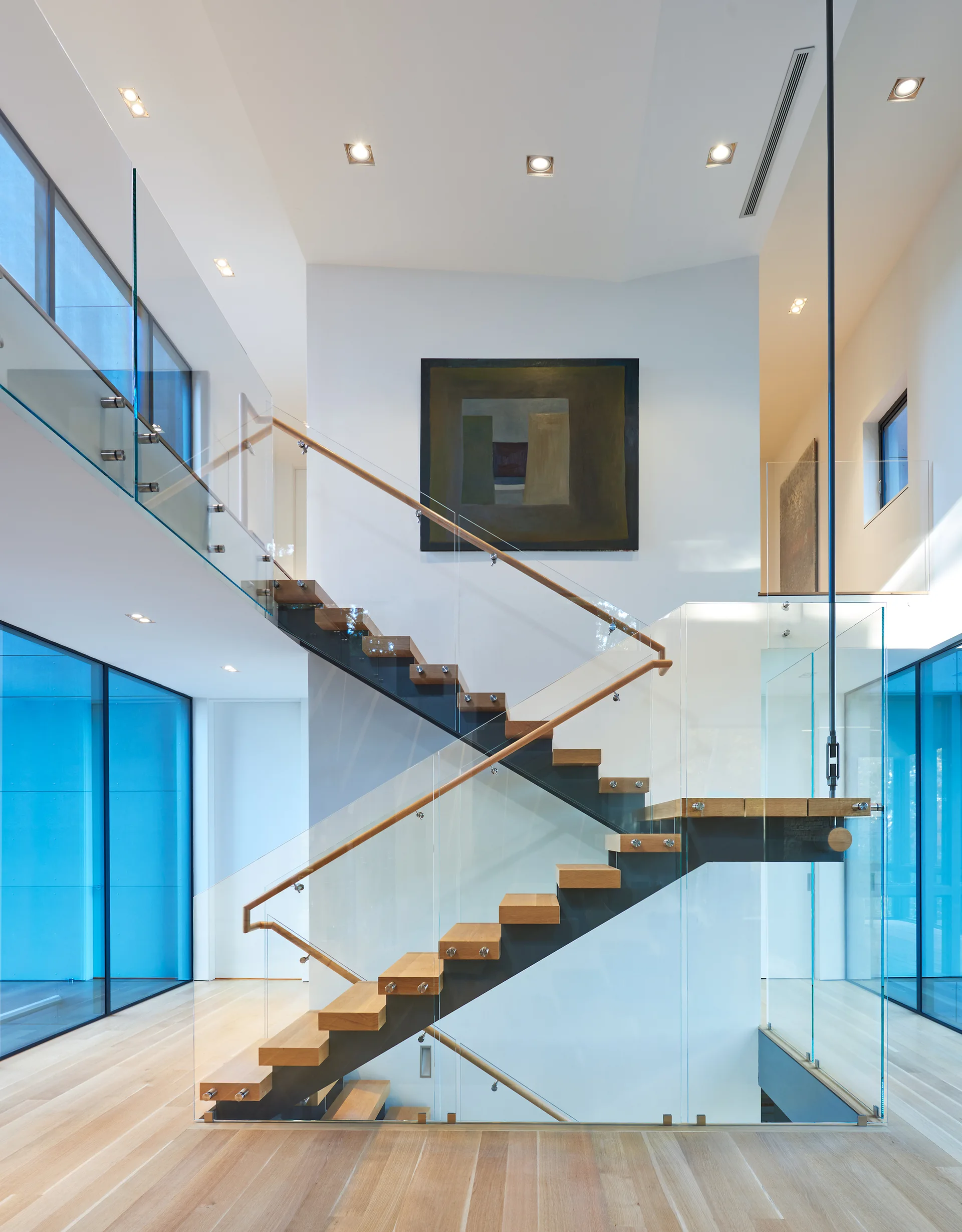 A modern floating staircase with light wood treads and clear glass railings sits against a white wall with an abstract painting.